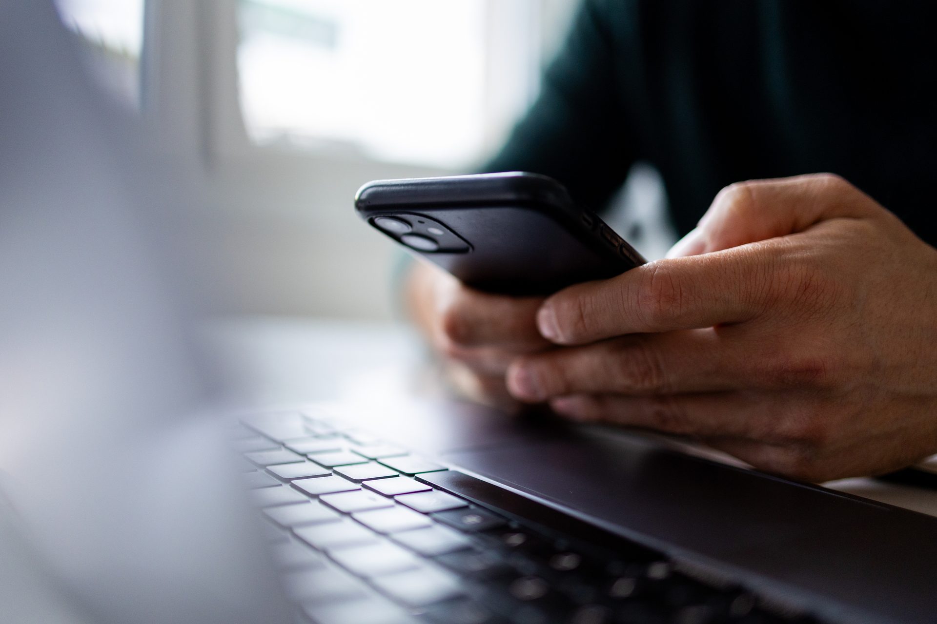 Close-up of a person's hands holding a black smartphone over a laptop keyboard.