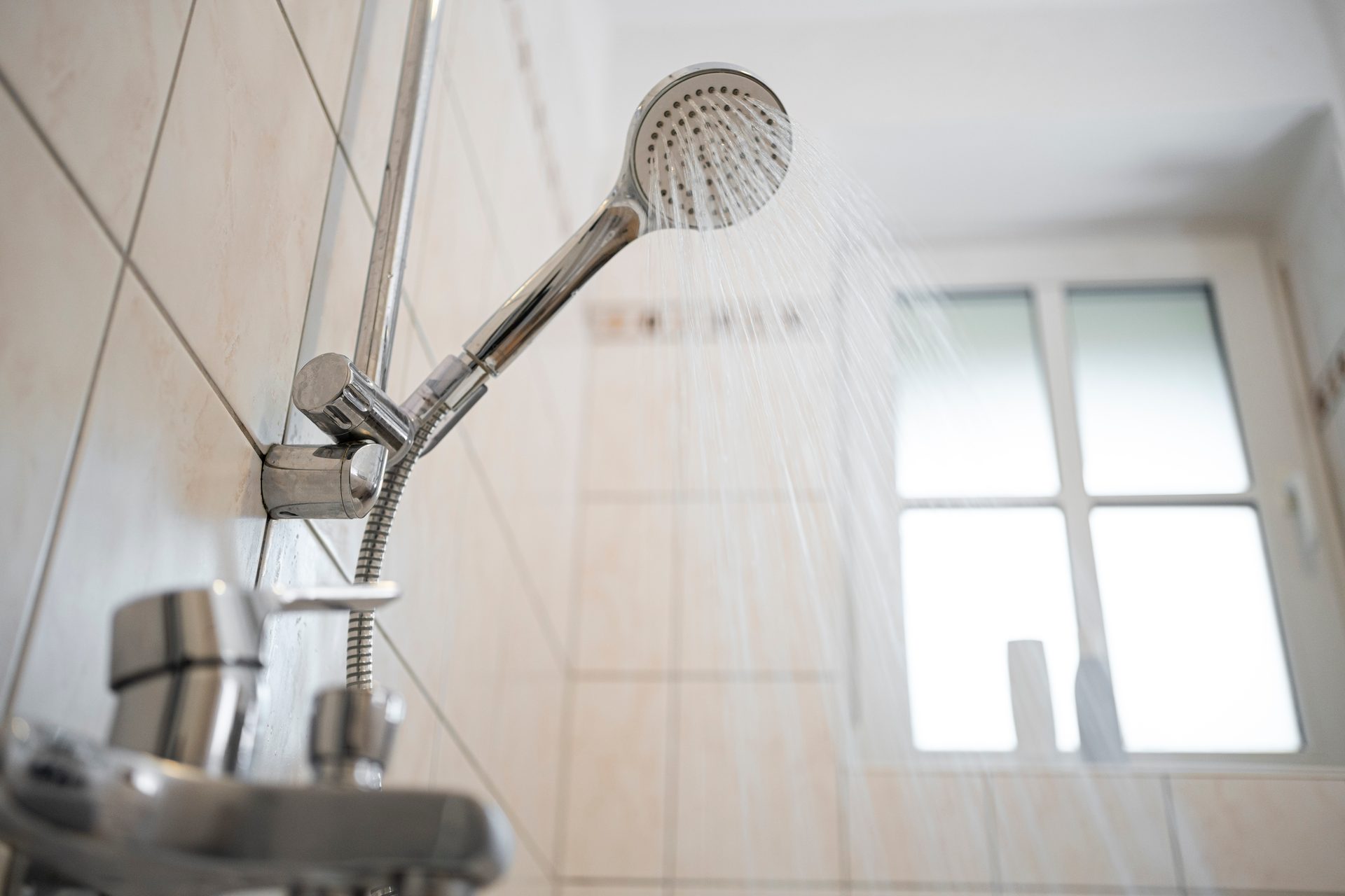 Close-up of a chrome shower head spraying water against a tiled bathroom wall and a bright window in the background.