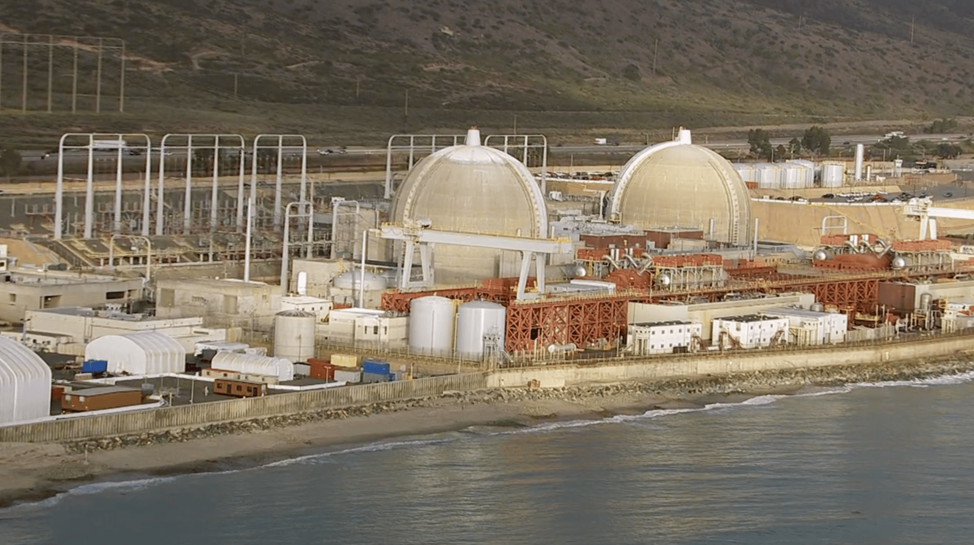 Nuclear power plant with two domes by the ocean, hills in the background.