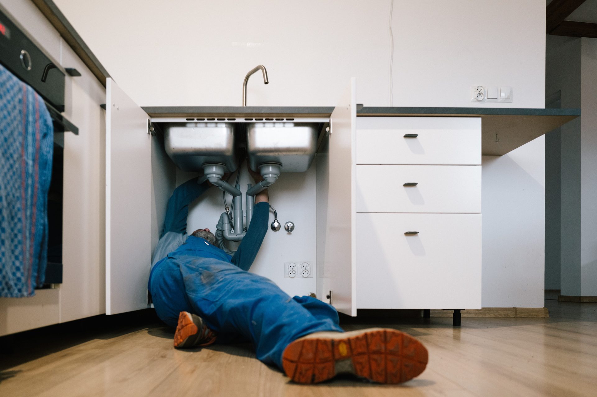 Man in blue overalls lies on his back fixing pipes under a double kitchen sink in a modern kitchen.