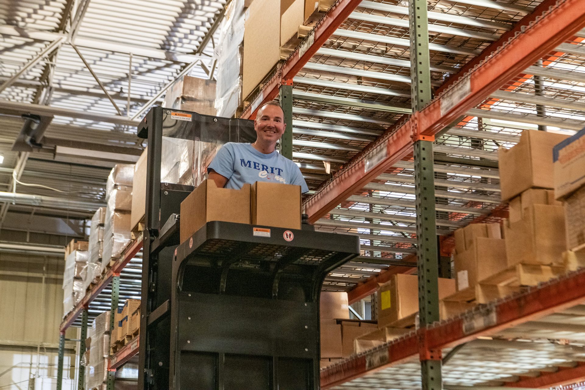 Smiling man on a forklift in a warehouse with tall shelves of boxes.