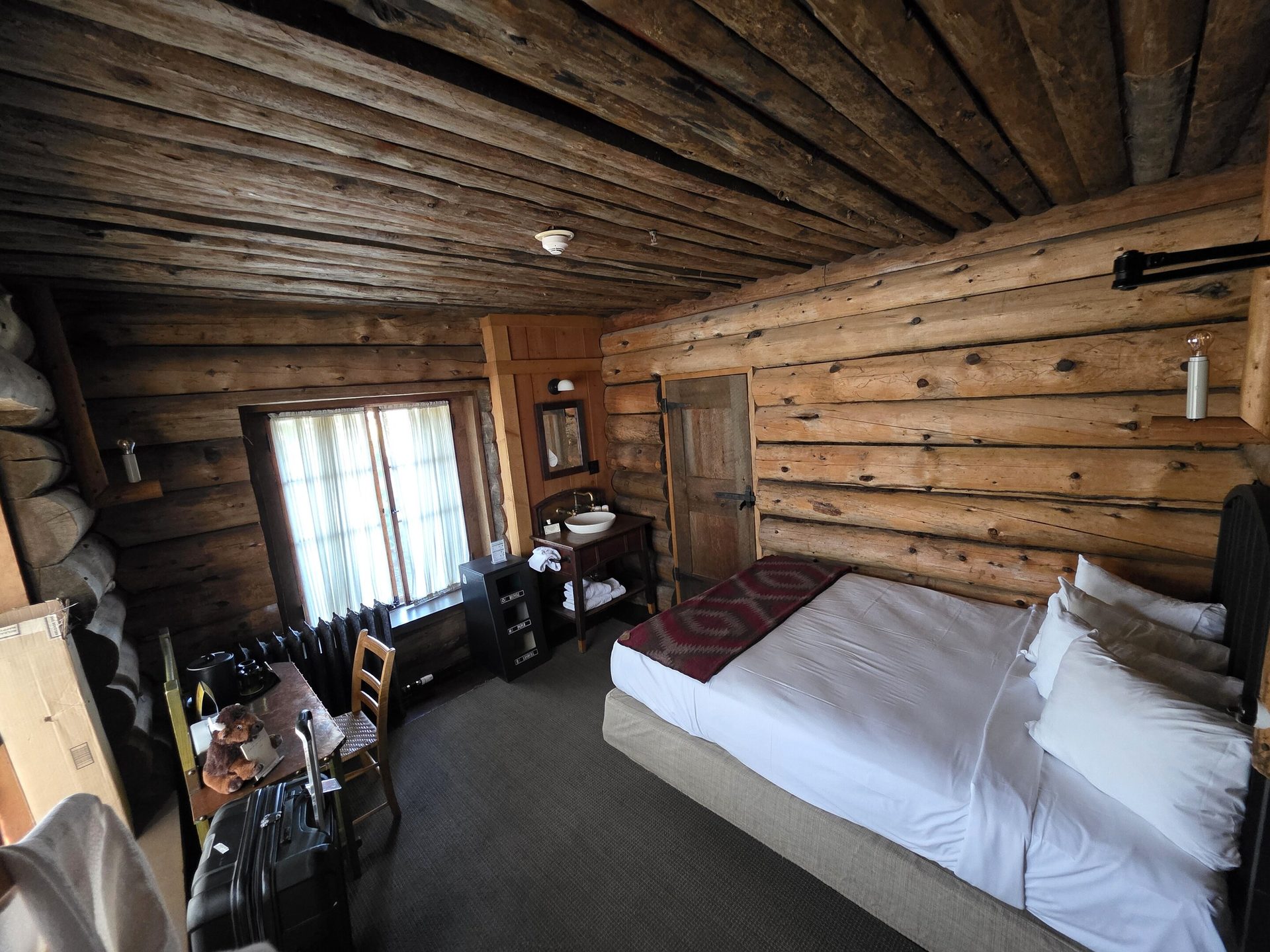 Rustic log cabin hotel room with a bed, window, desk, and sink, all in a wooden interior.