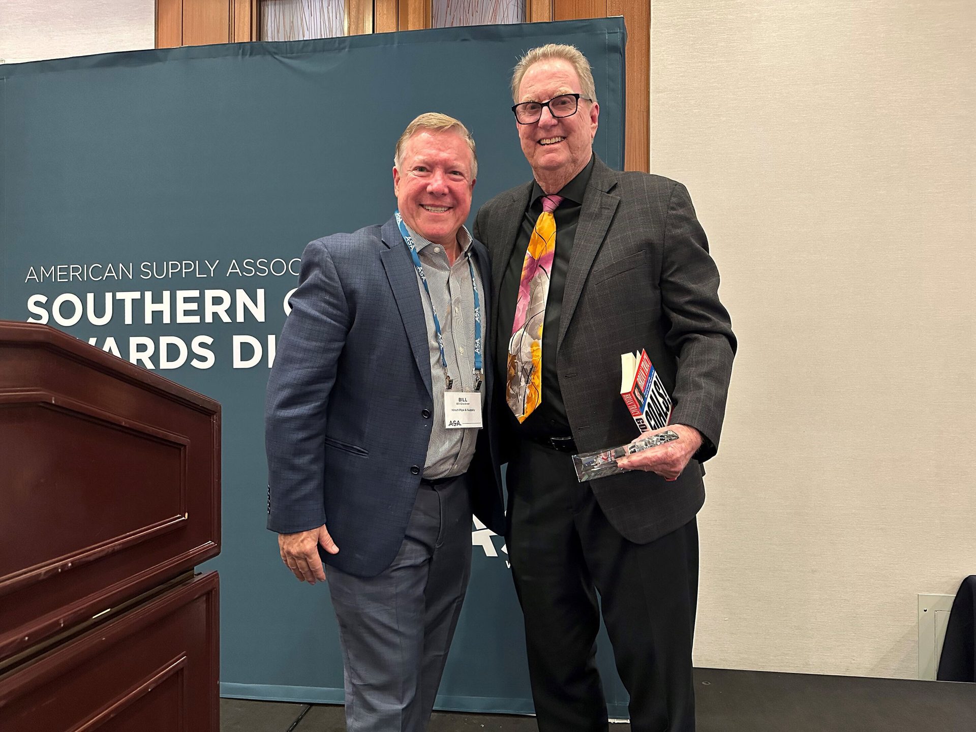 Two smiling men, one holding a book and an award, in front of an American Supply Association banner.