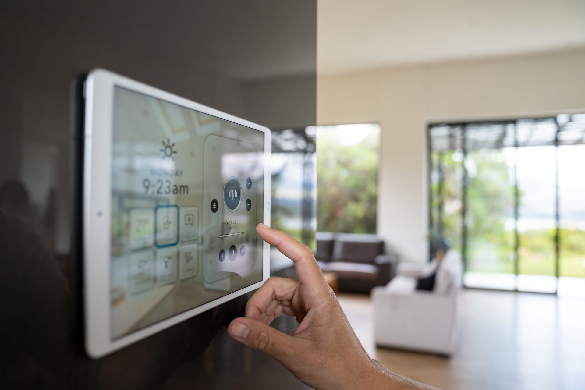 Hand operating a smart home control tablet mounted on a wall in a modern living room.