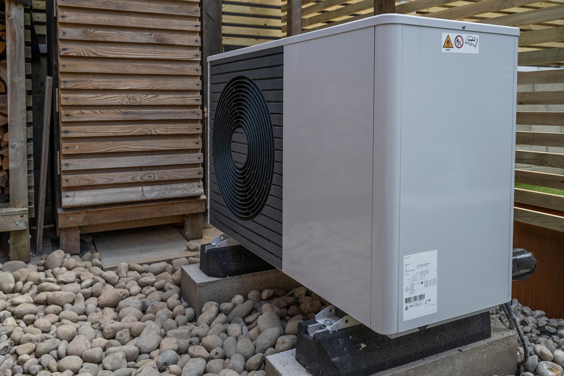 Gray outdoor heat pump unit with a black fan, on concrete blocks amidst pebbles and wooden slats.