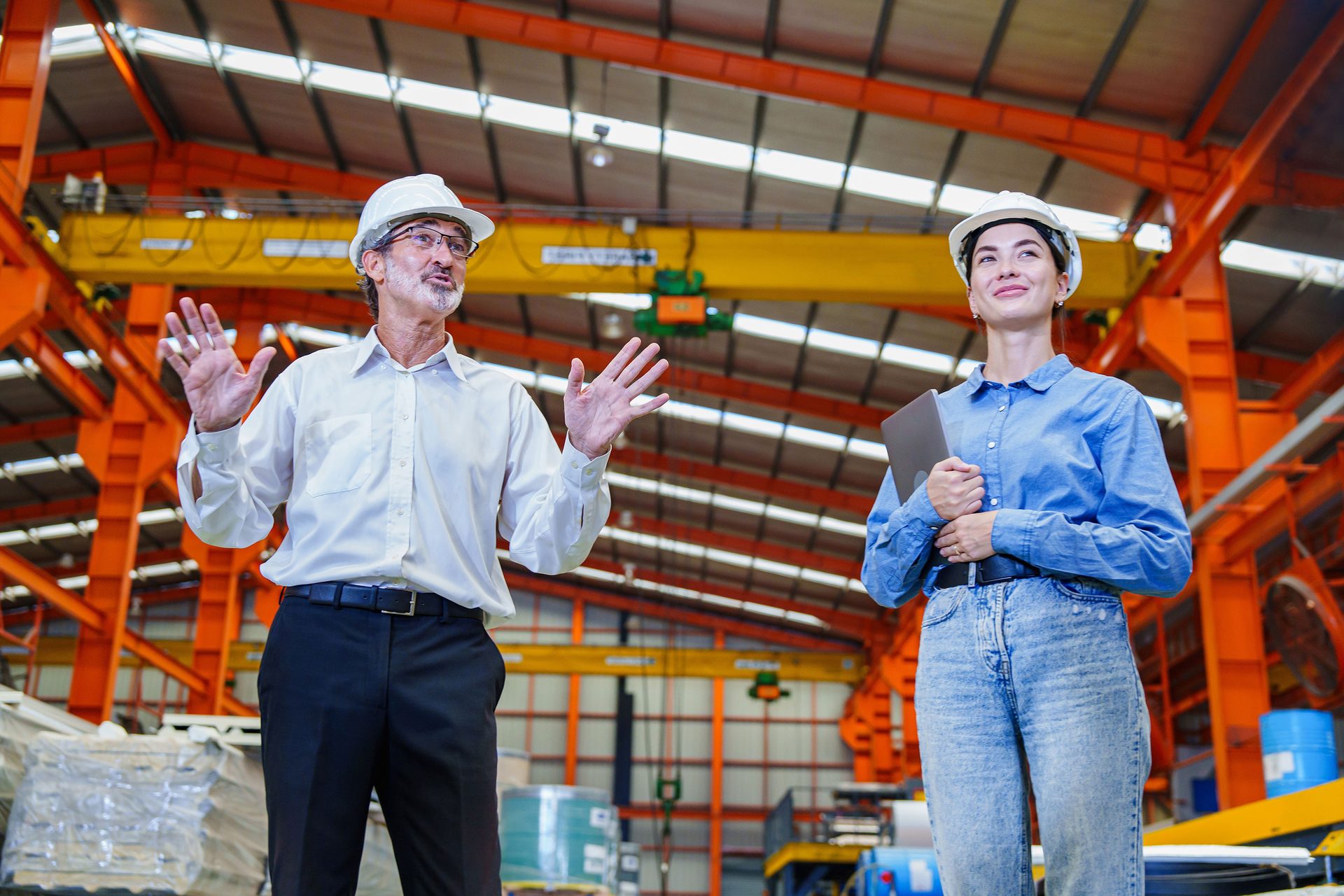 Two engineers in hard hats inspect a factory, man gesturing, woman holding a tablet.