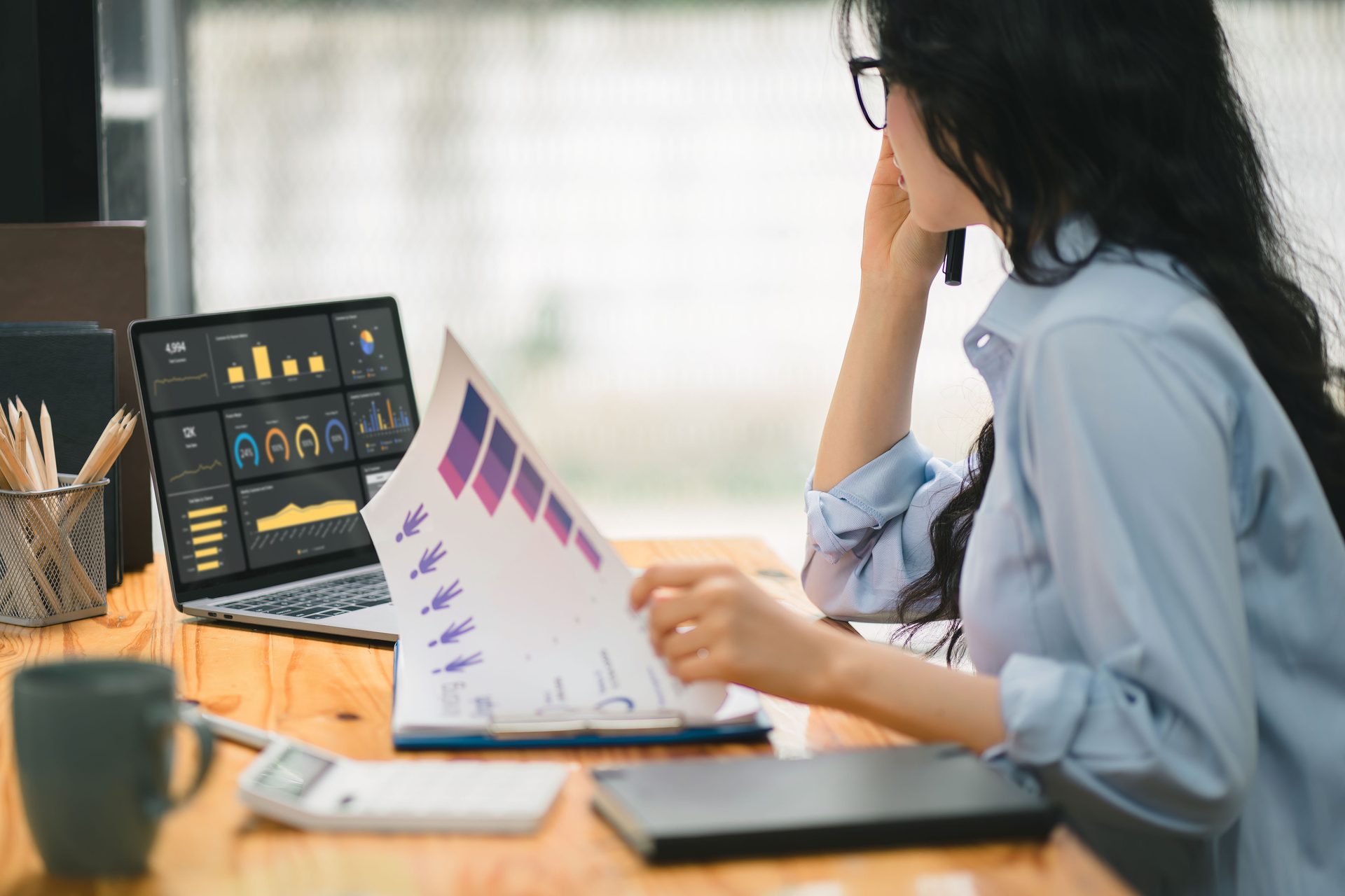 Person analyzing business data on a laptop and documents with charts at a desk.