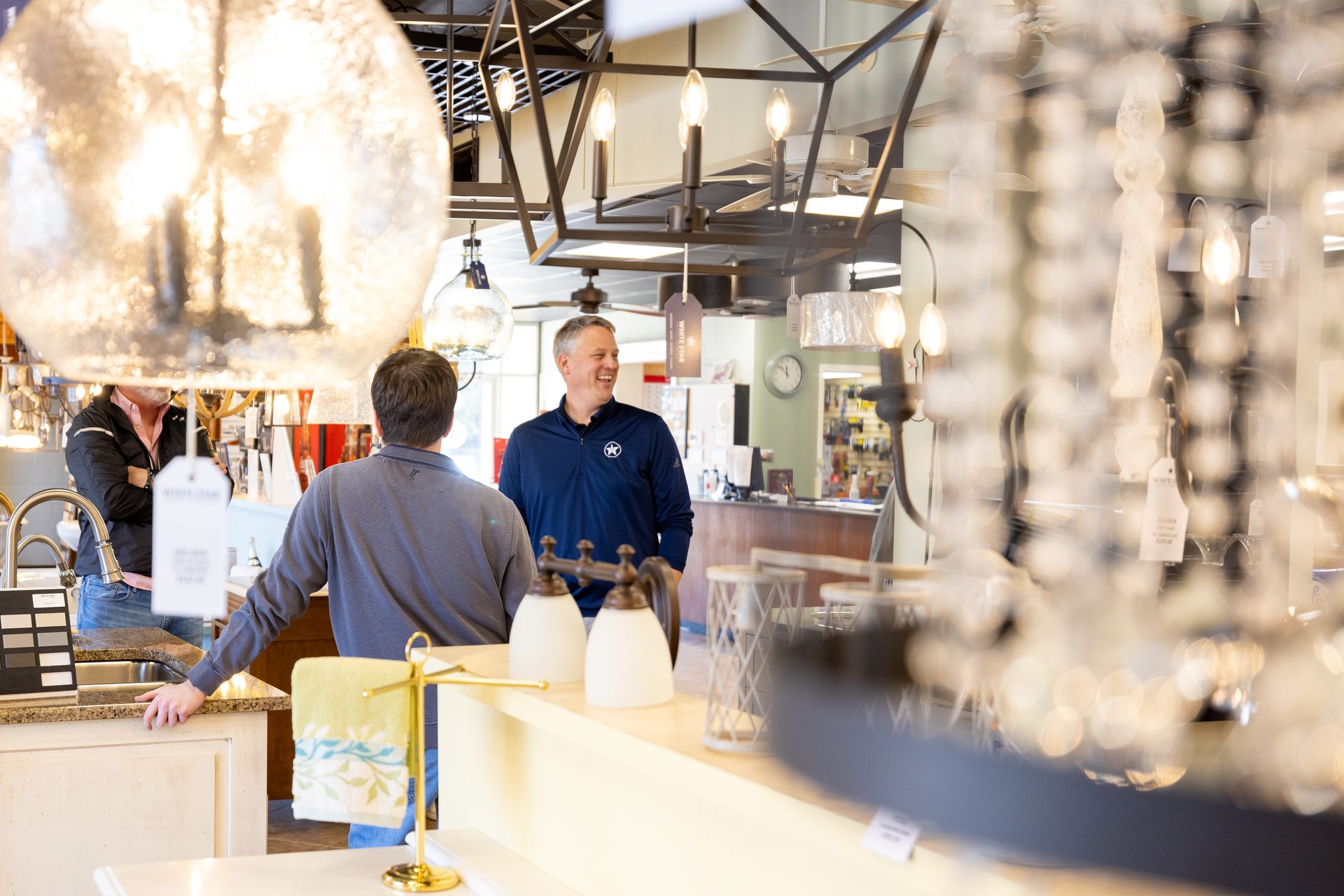Man in a blue shirt smiling while talking to another man in a lighting and home decor store.