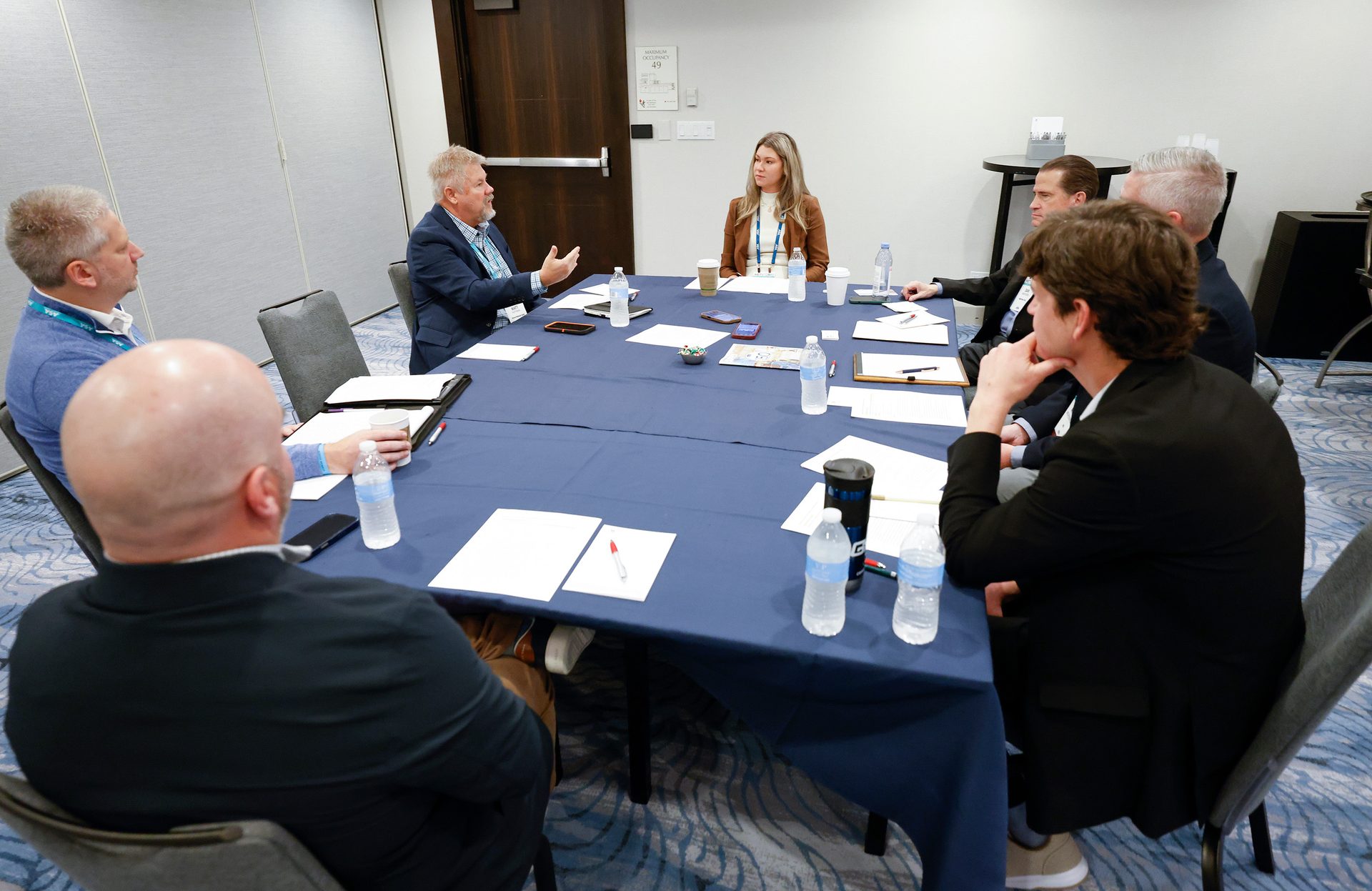 Eight people gather around a blue conference table, engaged in a meeting.