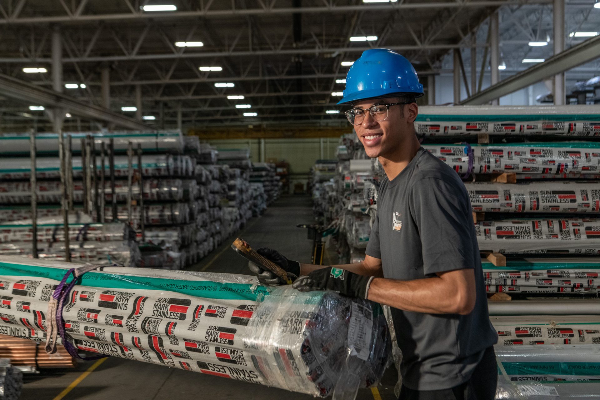 Smiling worker in hard hat handles wrapped stainless steel pipes in a warehouse.