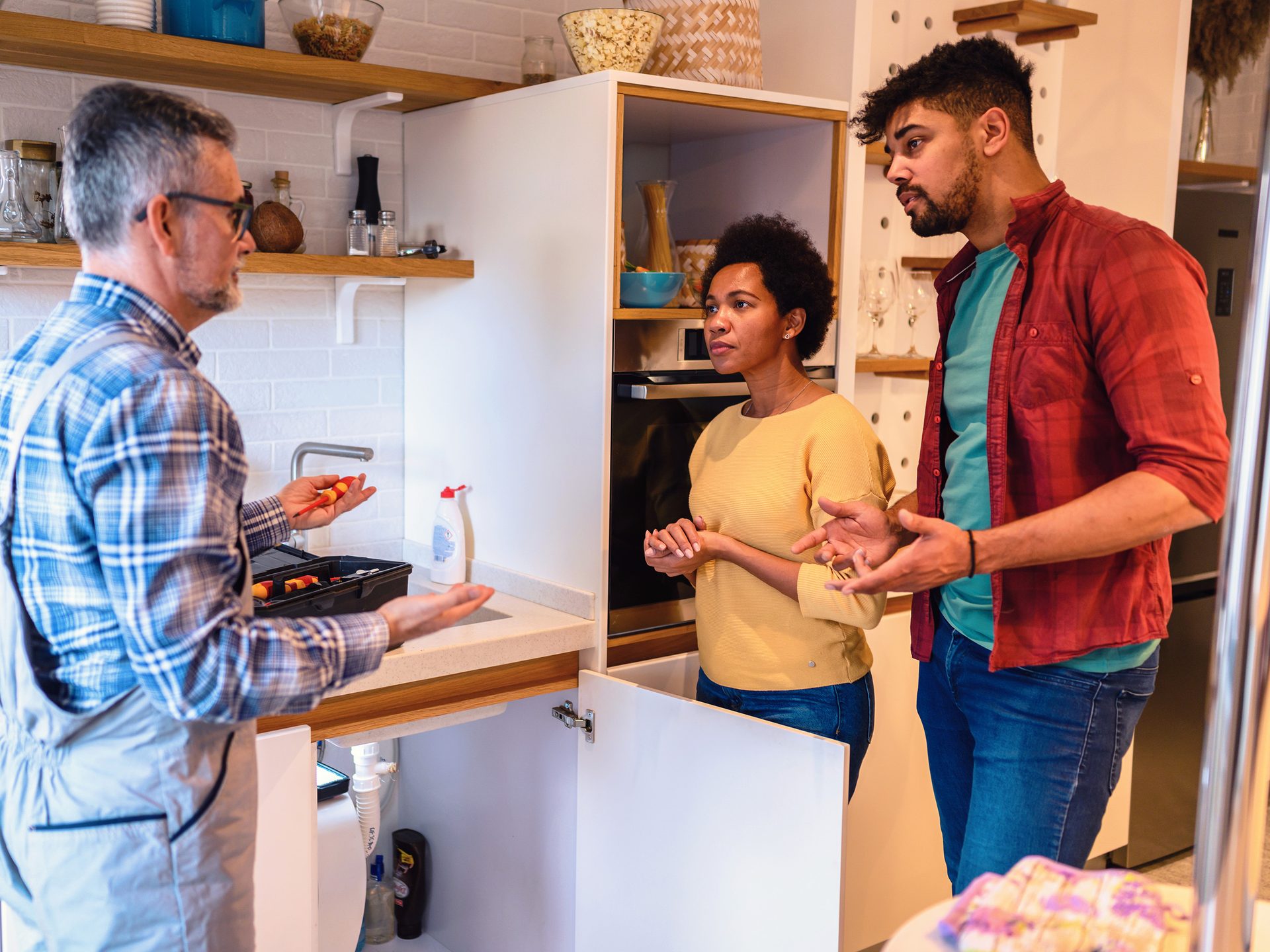 A plumber explains a repair issue to a concerned couple in their kitchen, standing near an open cabinet under the sink.