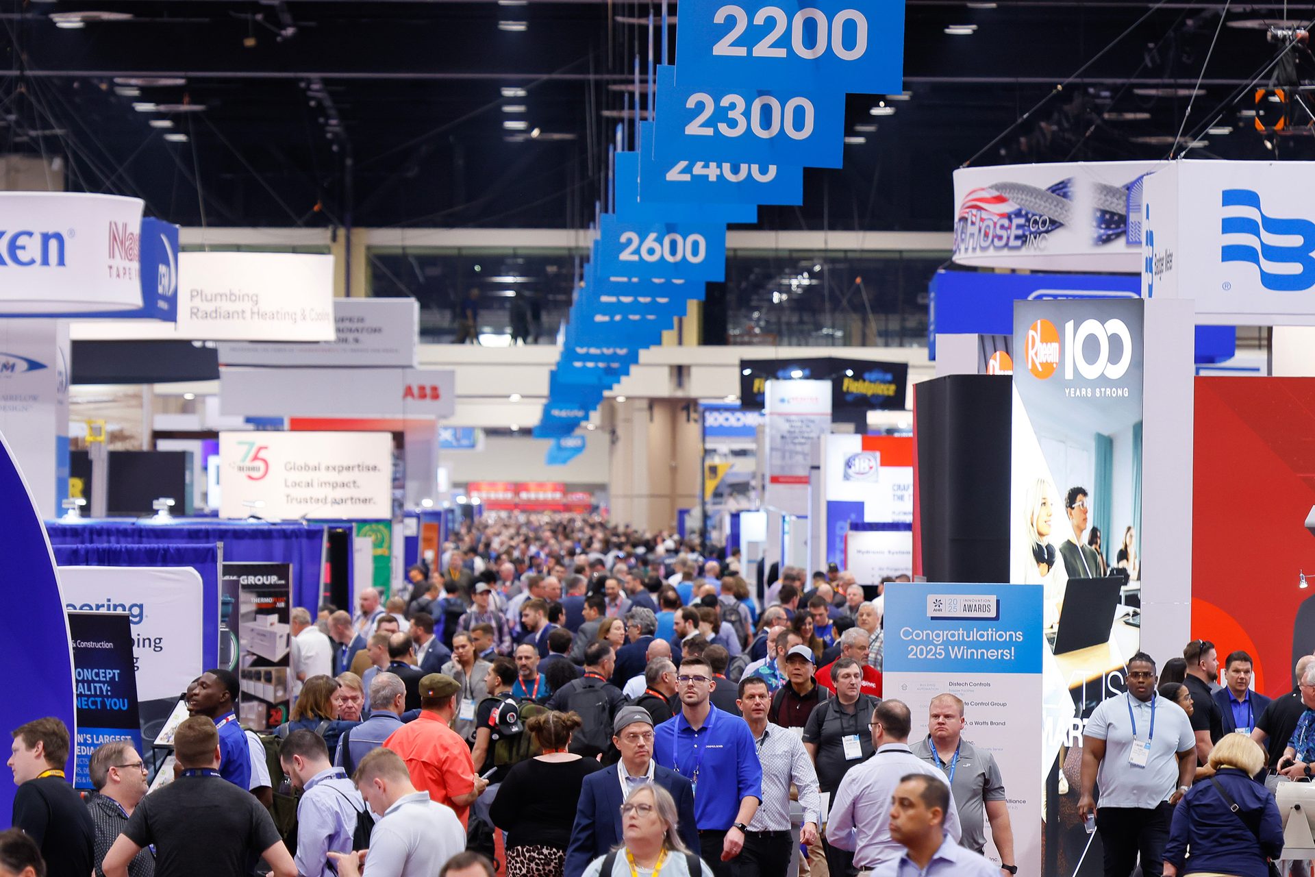 Crowded trade show with attendees, booths, and overhead numbered section signs.