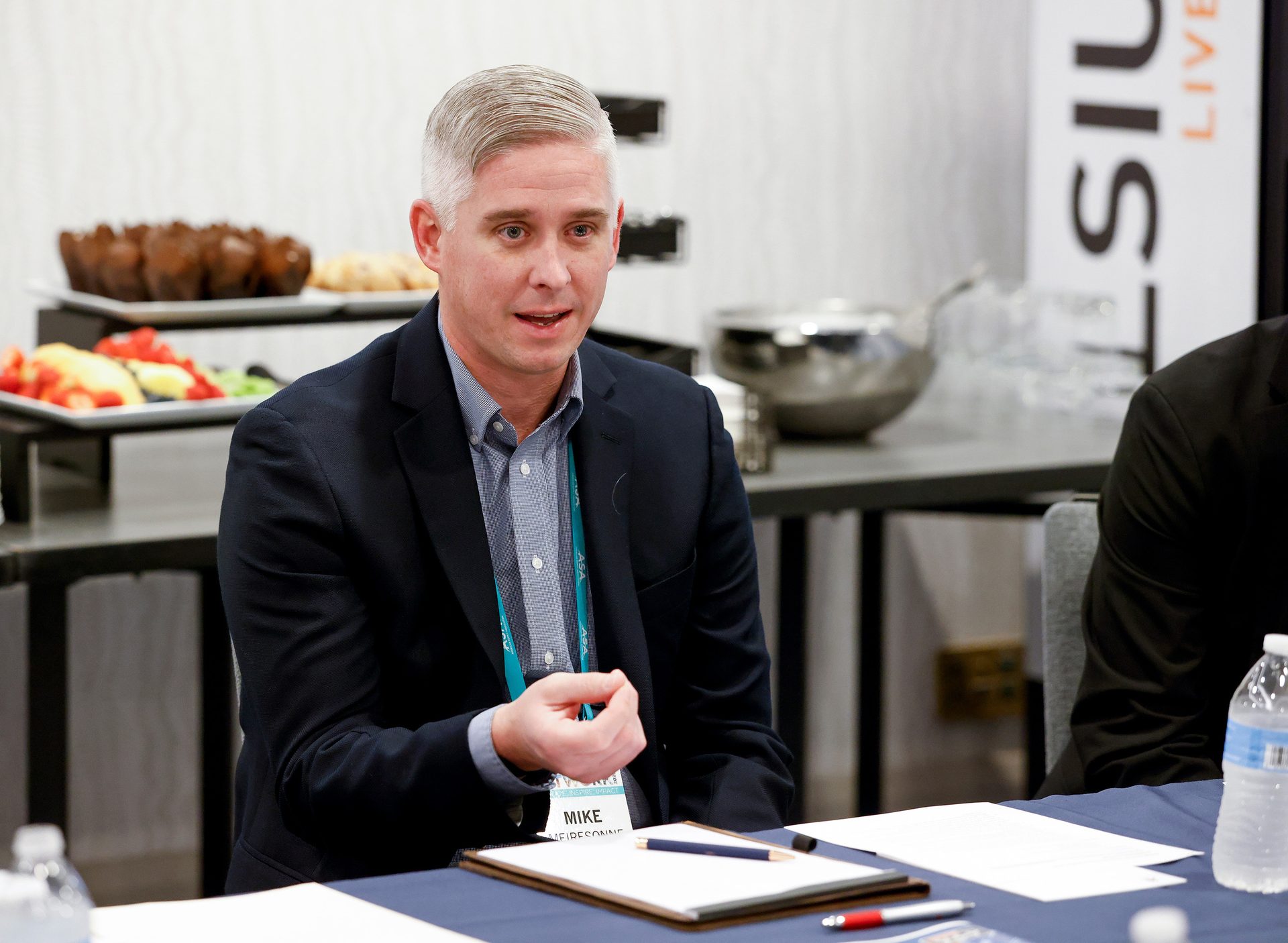 A man named Mike in a suit speaks at a conference table, with food and drinks behind him.