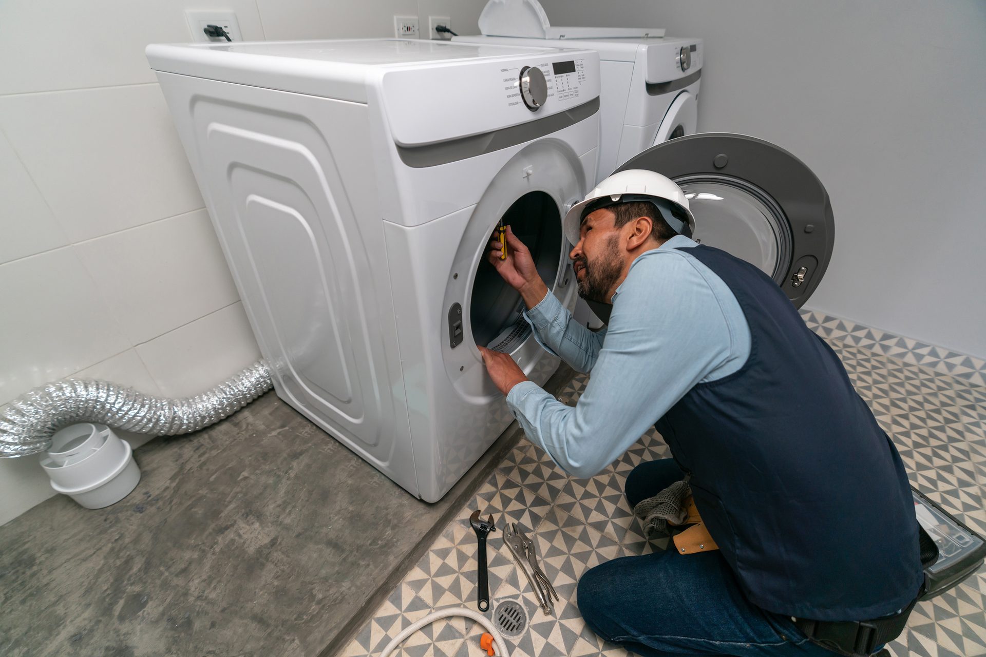 A man in a hard hat and work clothes repairing a front-load washing machine with a screwdriver.
