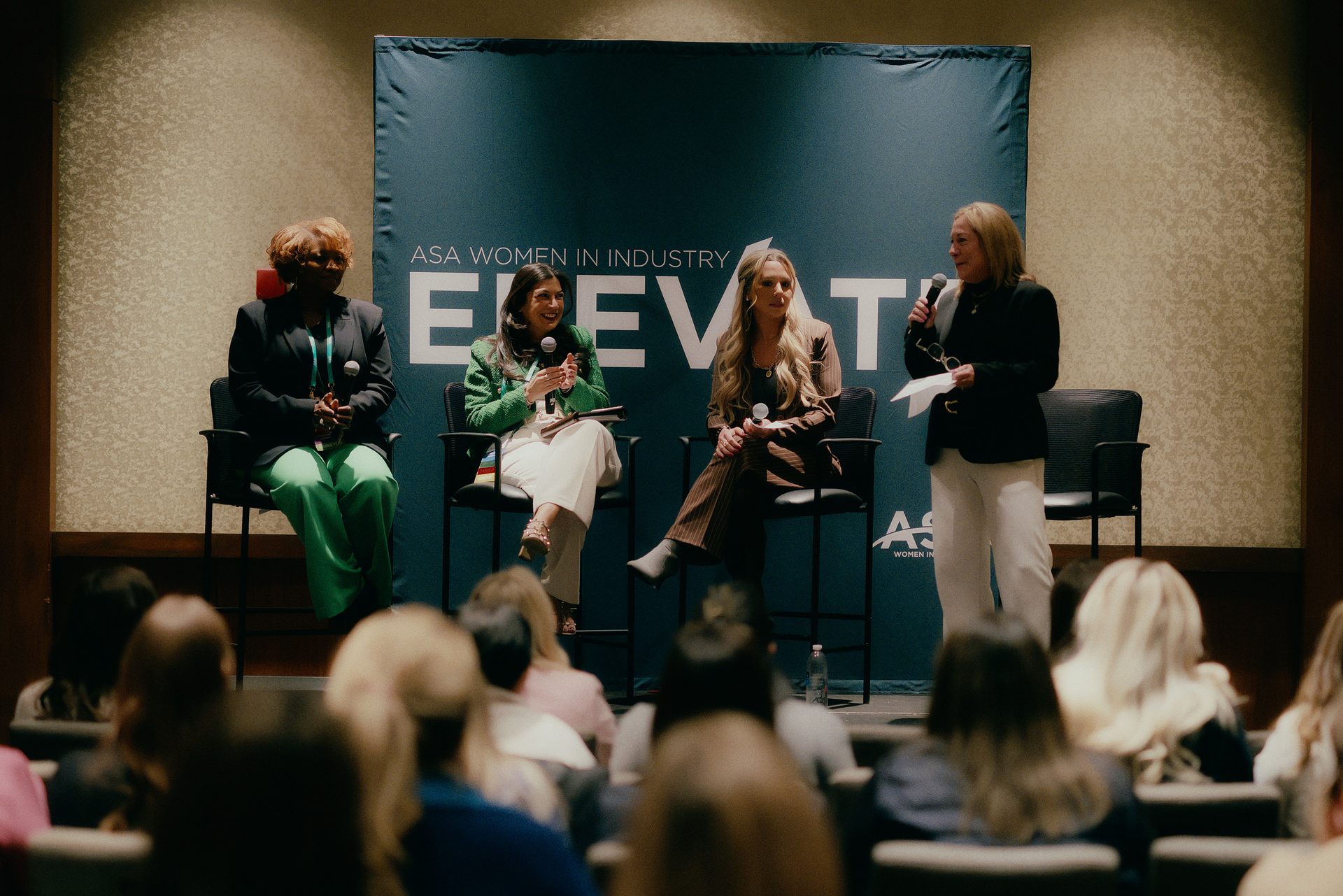 Four women, including a panel of three seated and one standing speaker, participate in a conference.