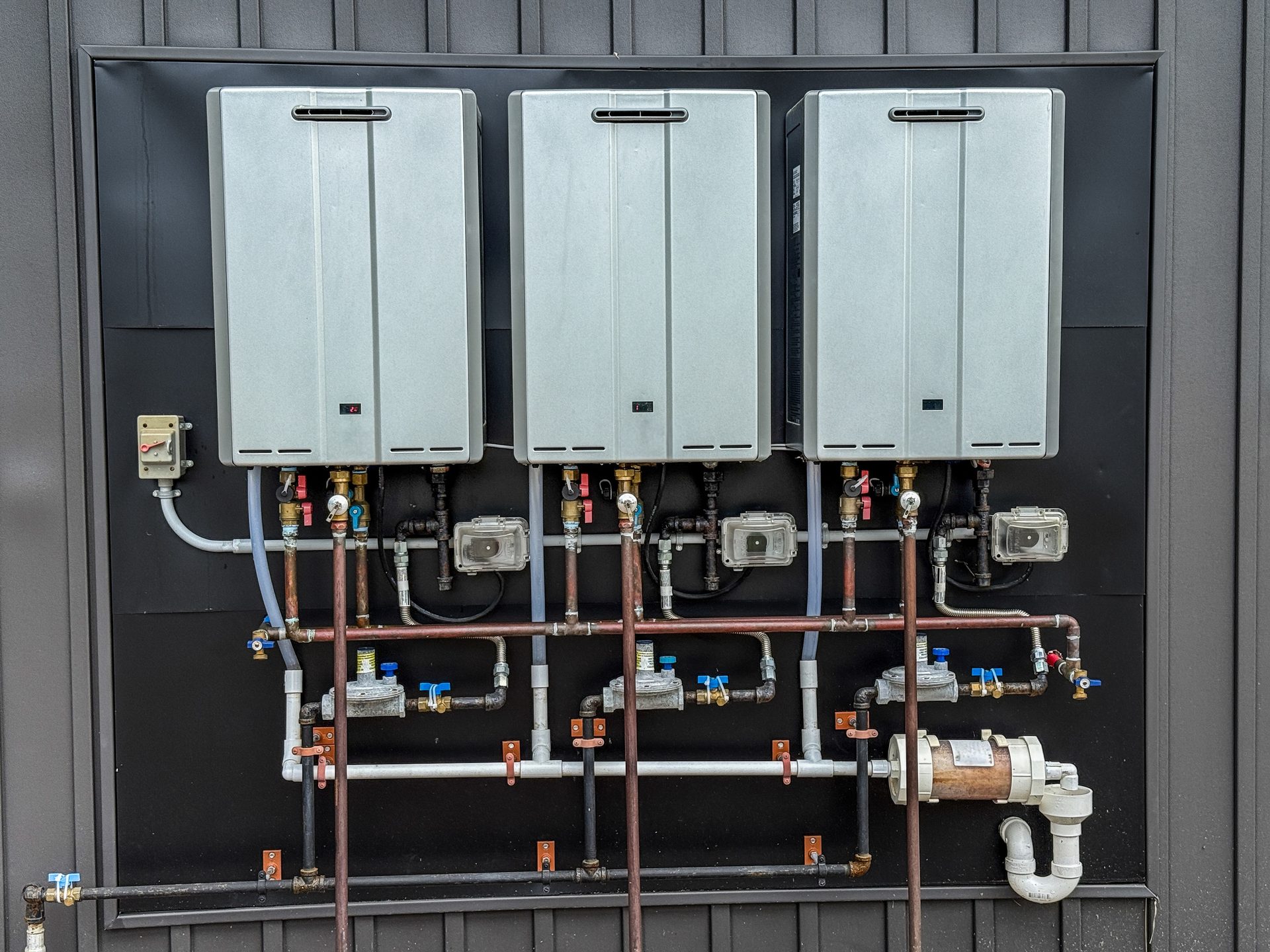 Three tankless water heaters with complex plumbing on a dark wall.