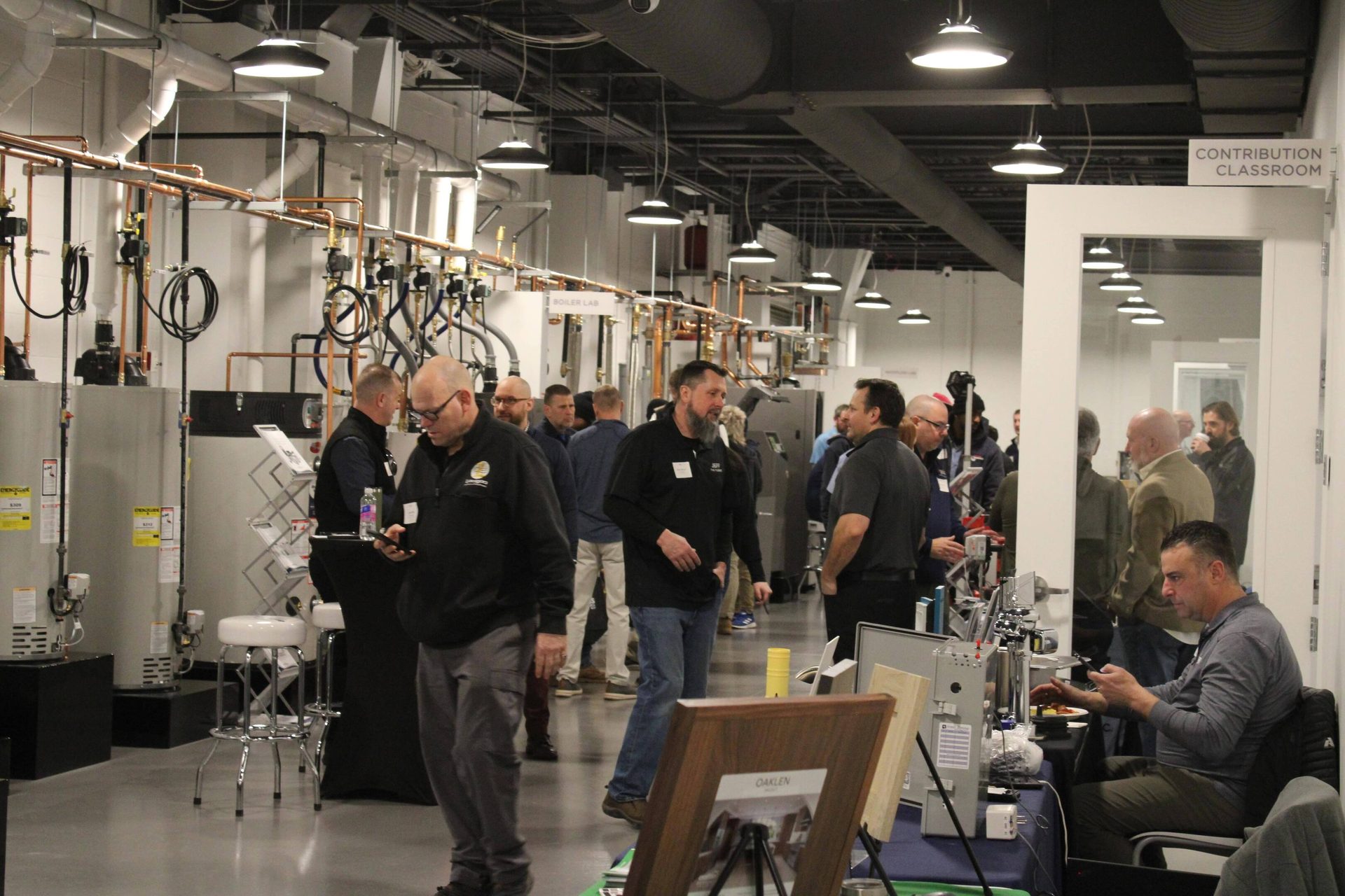 Men gather in a technical lab with boiler equipment and pipes, discussing and engaging with displays.