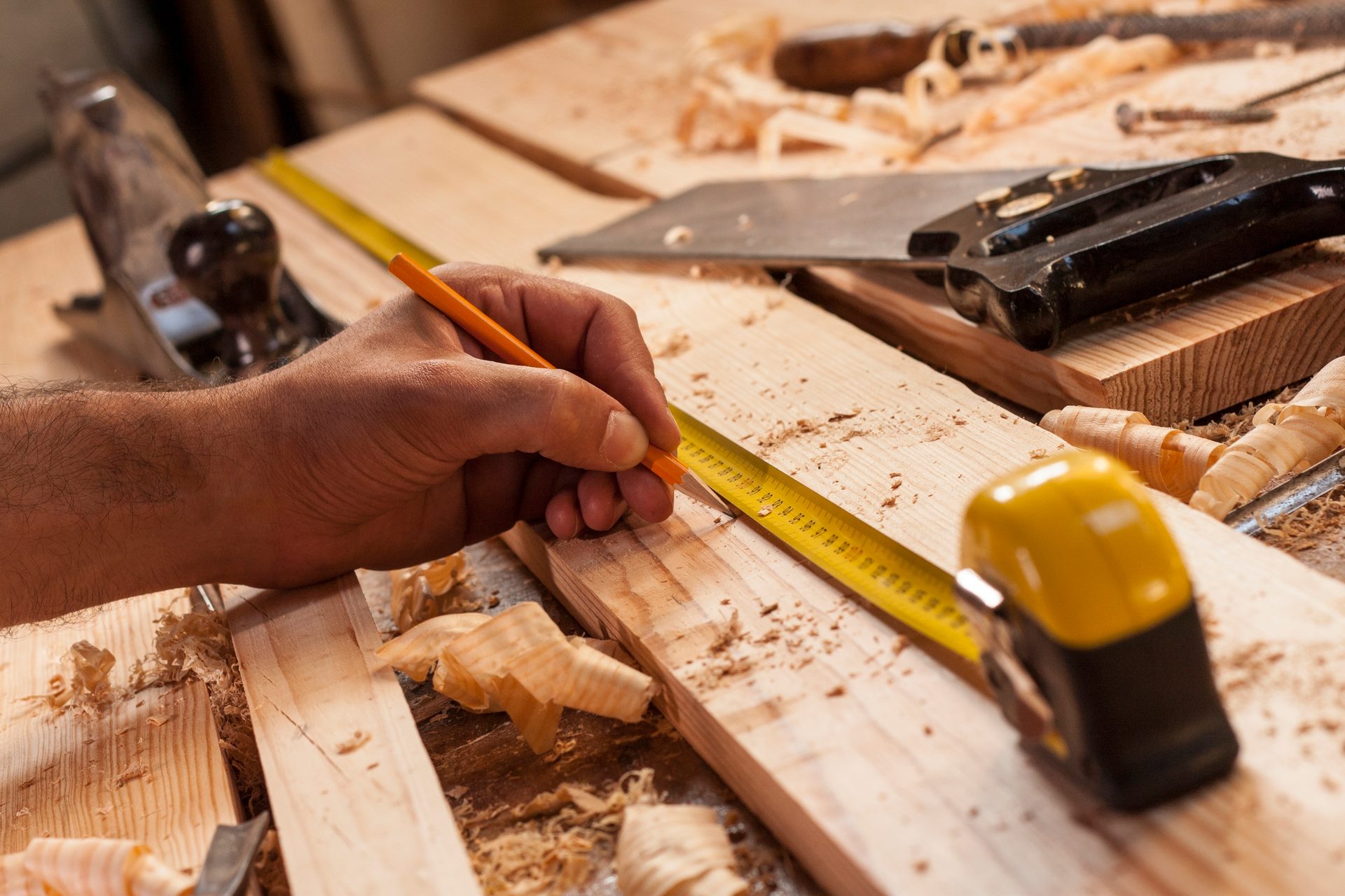 A carpenter's hand measures a wooden plank with a pencil and tape, surrounded by wood shavings.