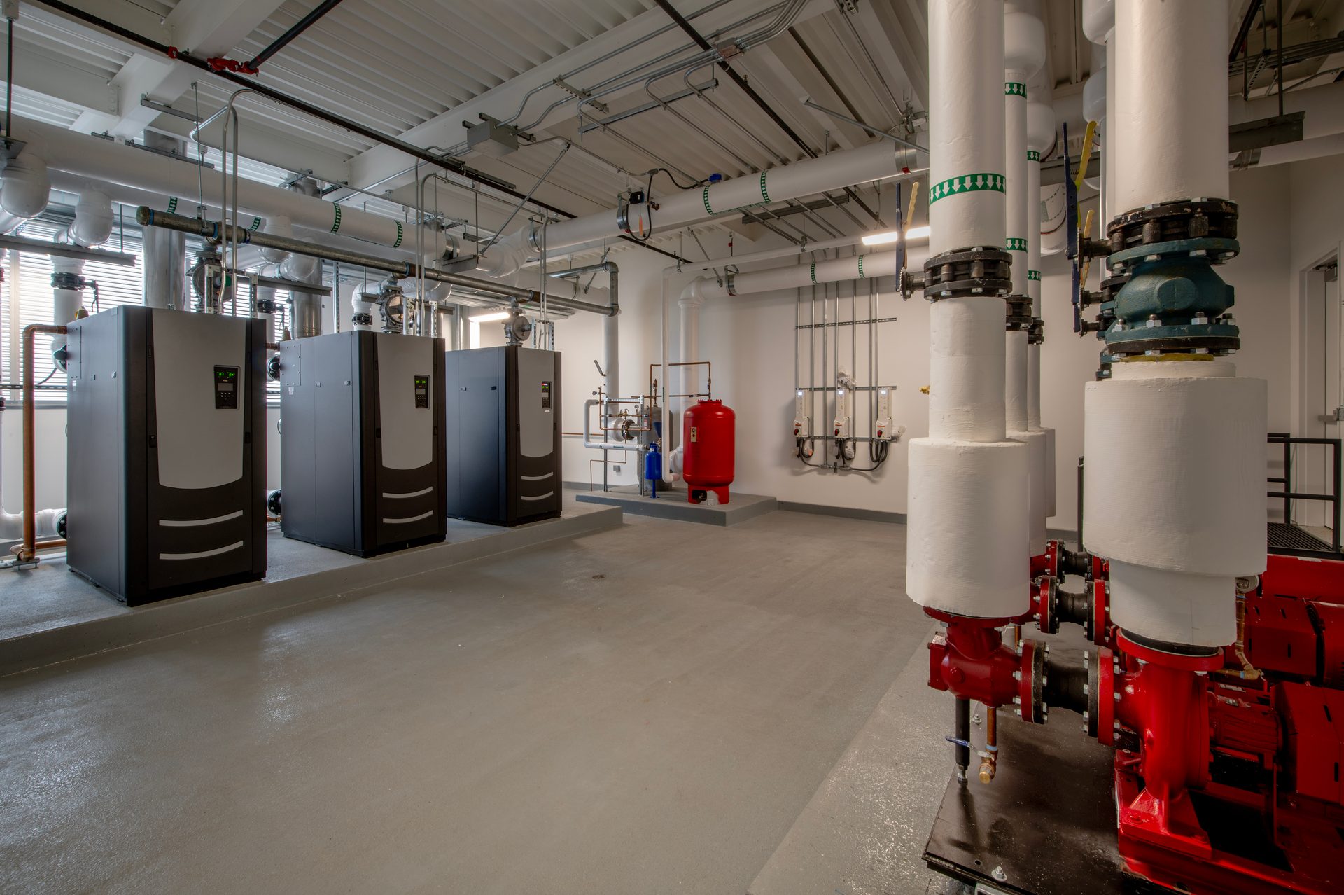 Modern boiler room with large heaters, red expansion tank, and complex piping system.
