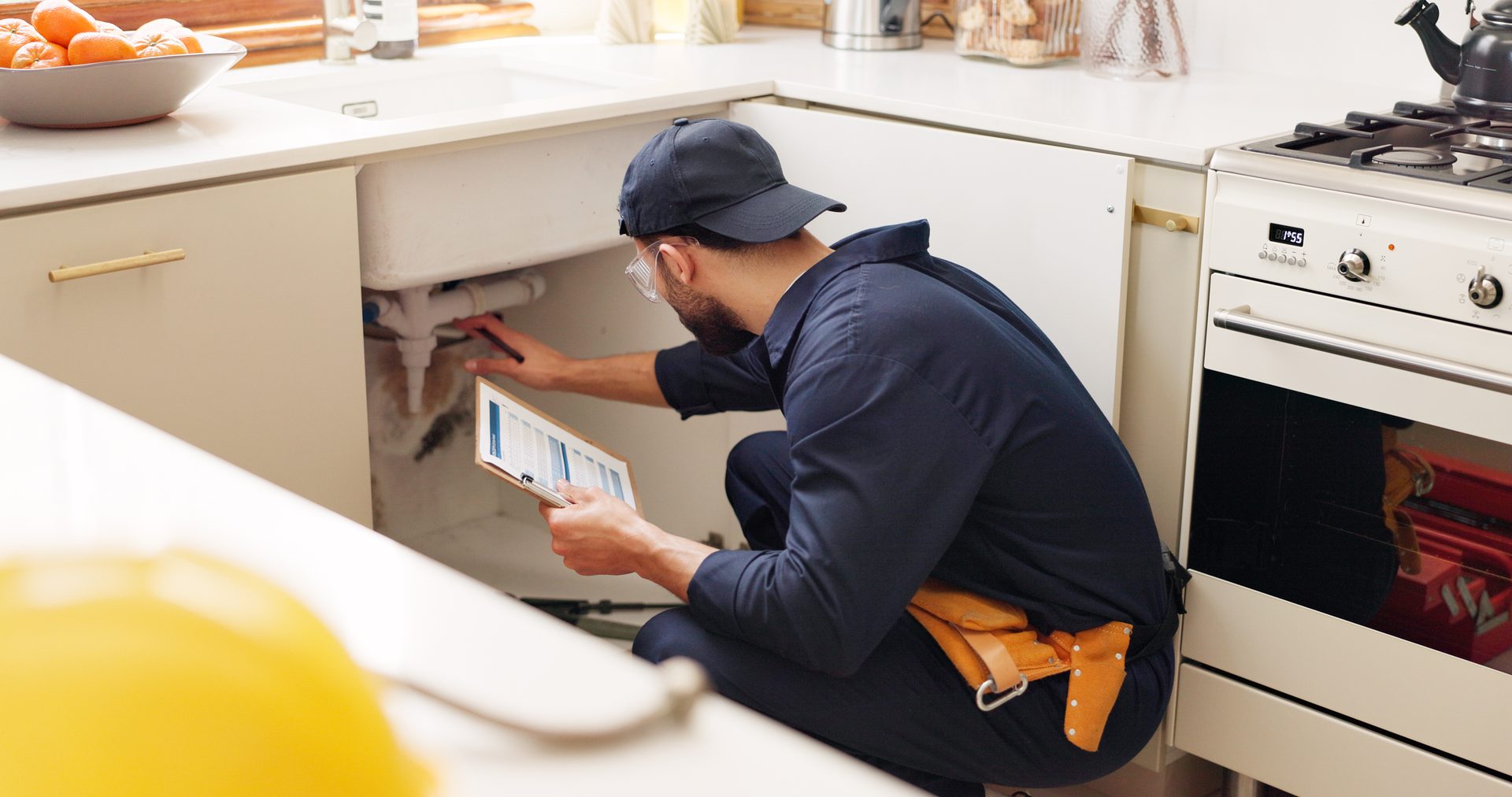 A plumber in a cap and safety glasses inspects pipes under a kitchen sink while holding a tablet.