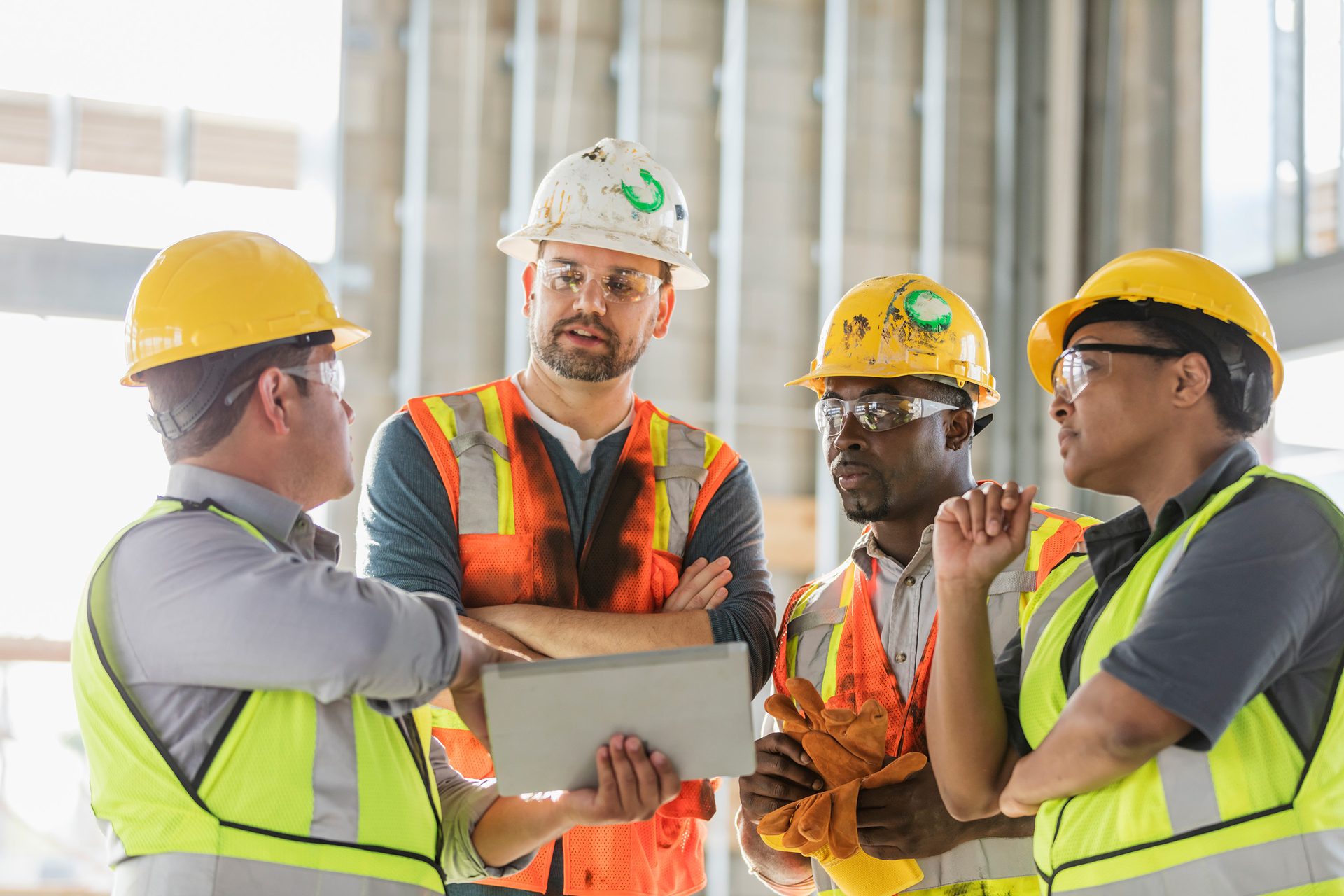 Four construction workers in hard hats and safety vests discussing plans on a tablet at a job site.