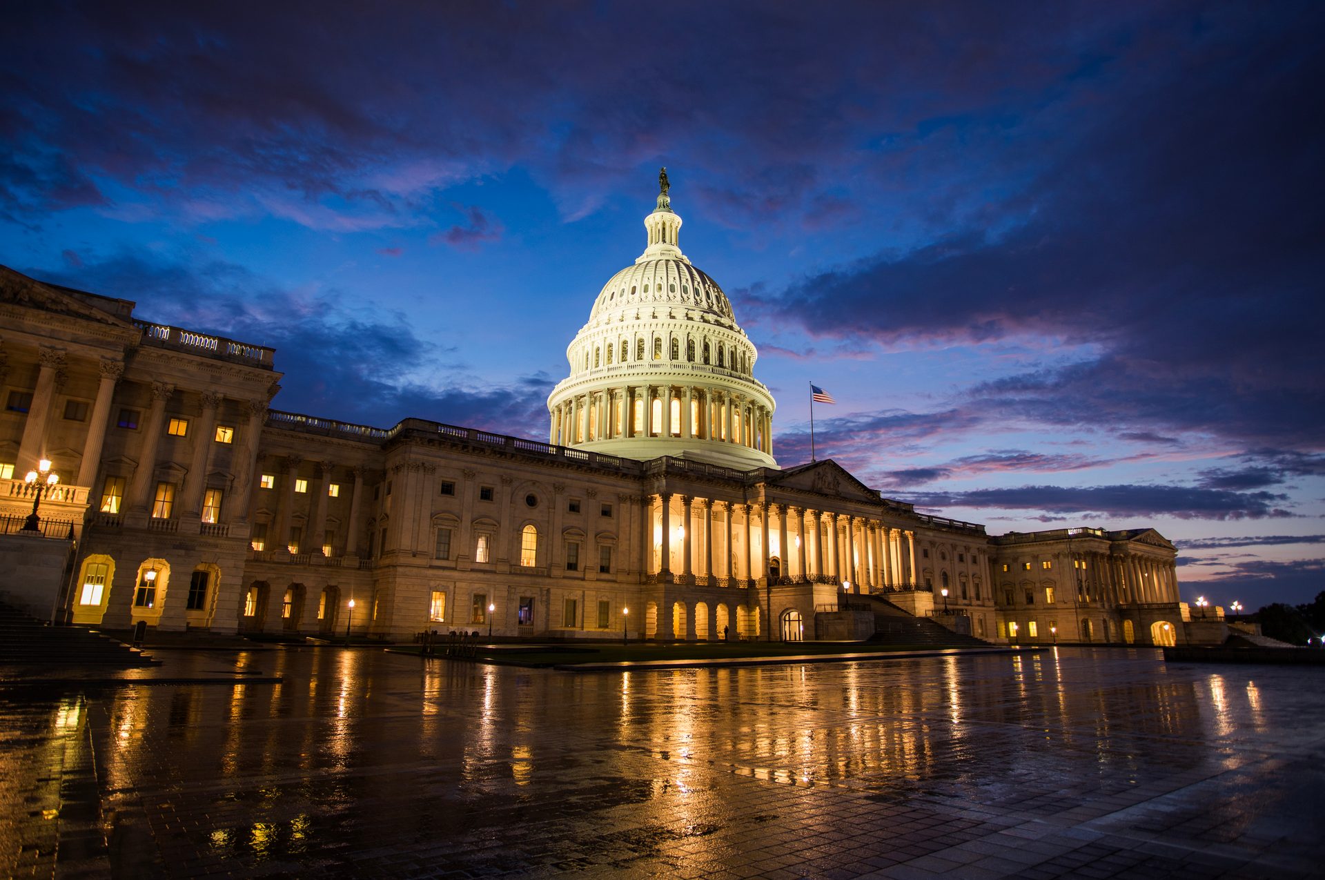 Illuminated US Capitol building at night with dramatic clouds and reflections on wet ground.