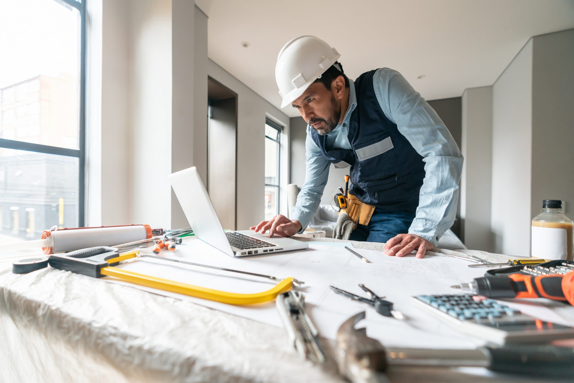 Construction worker in hard hat and vest using a laptop on a table with blueprints and tools.