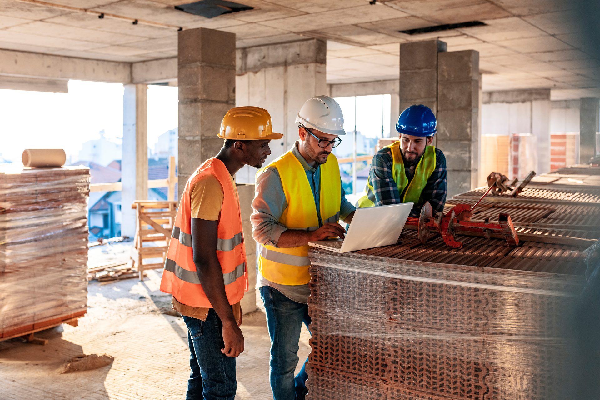 Three workers in hard hats and safety vests review a laptop on a construction site.