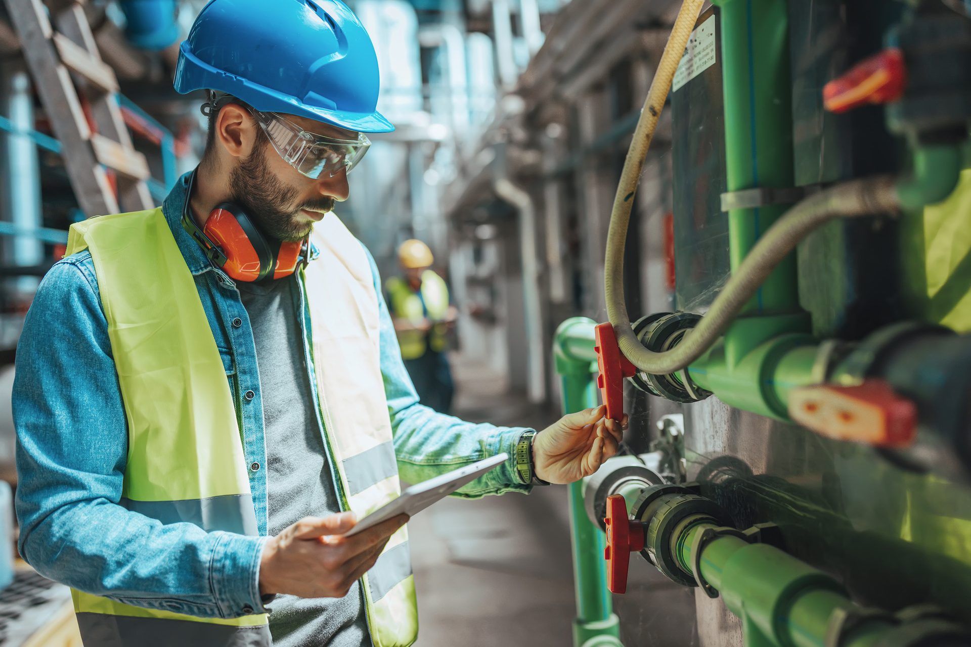 Engineer in safety gear inspects industrial piping and valves with a tablet.