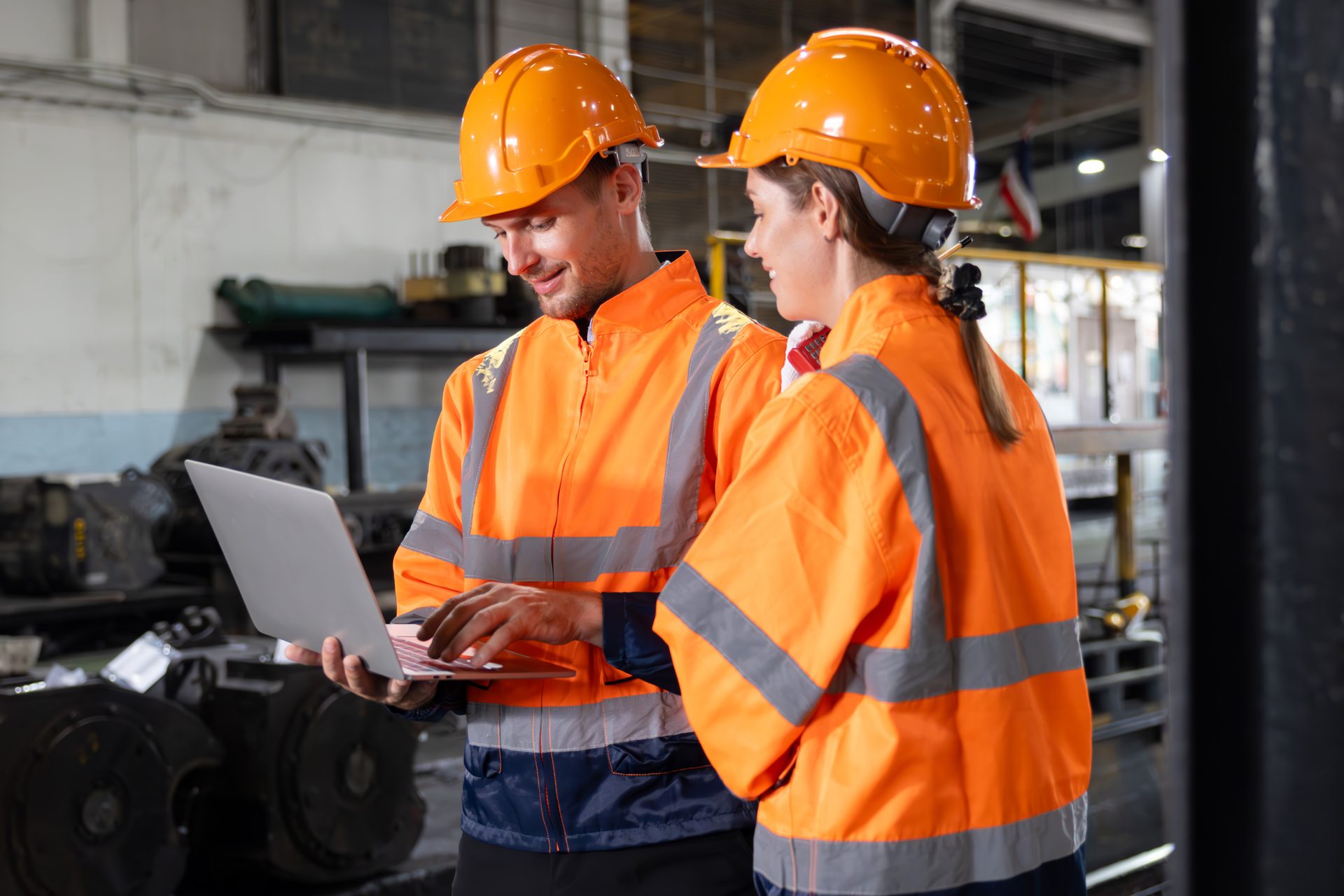 Two workers in orange safety vests and hard hats in a factory, using a laptop.