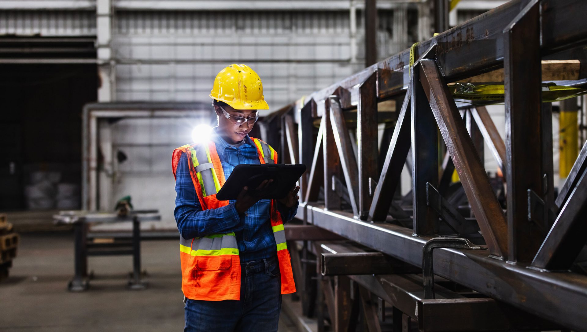 A woman in a hard hat and safety vest inspects a metal structure with a tablet.