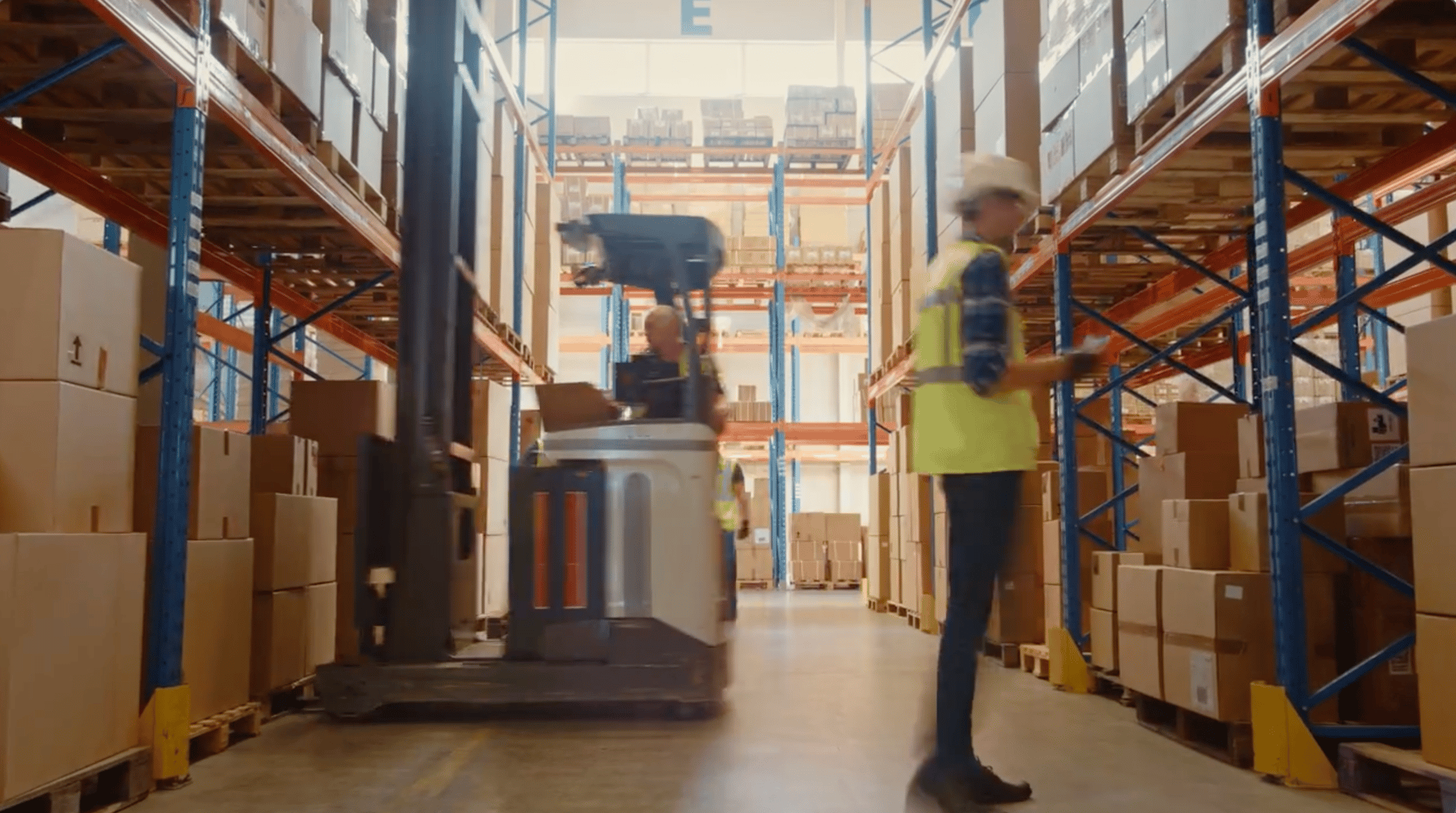 A worker with a tablet and a forklift operator move through a busy warehouse aisle filled with boxes.