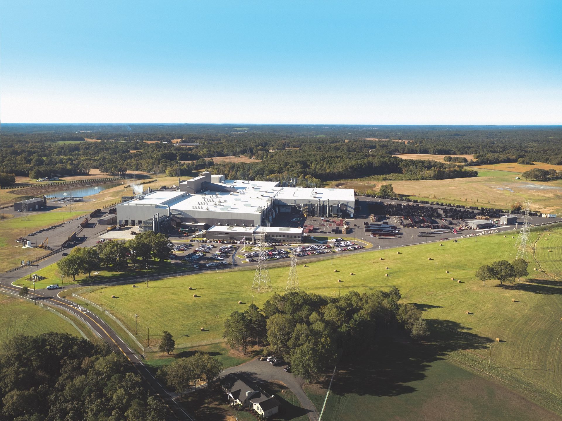 Aerial view of a large industrial plant with a white roof, surrounded by green fields and trees under a clear sky.