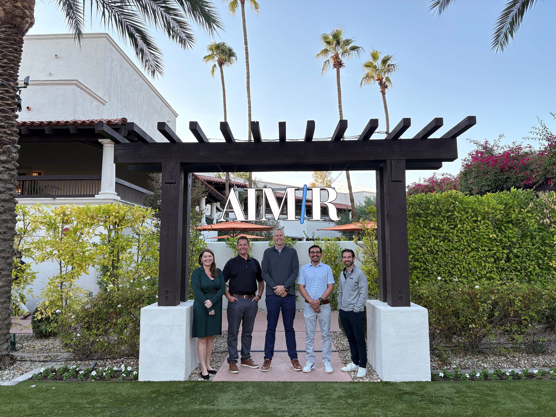 Five professionals stand under a wooden archway spelling "AIMR" with palm trees in the background.