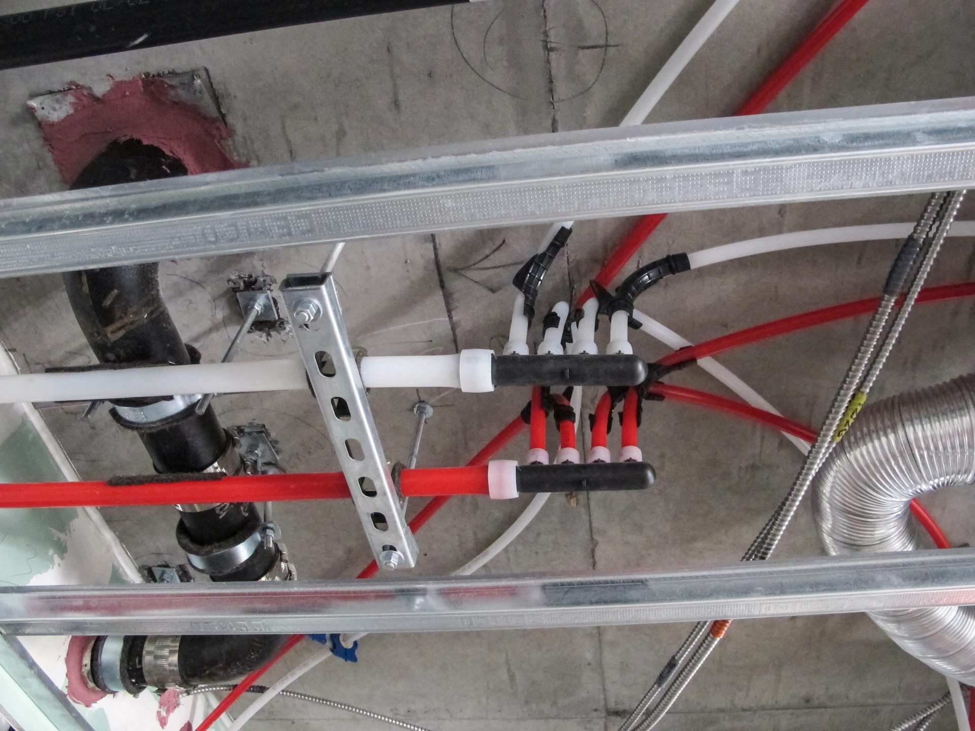 Complex network of plumbing pipes (PEX) and metal framing attached to a concrete ceiling, with HVAC ducting.