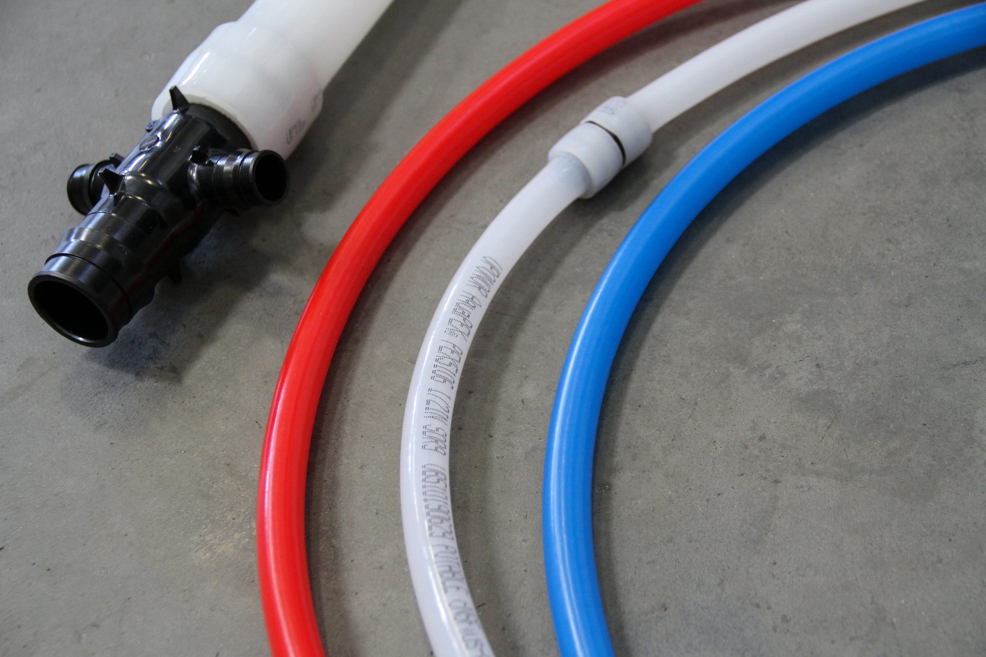 Close-up of red, white, and blue PEX plumbing pipes with a black multi-port fitting on a concrete background.