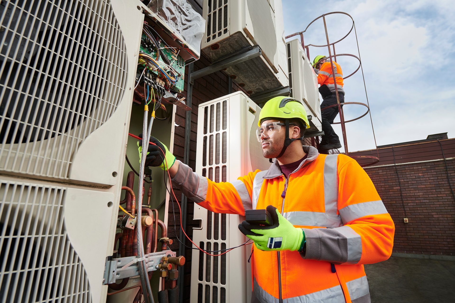 Technicians in safety gear servicing HVAC units on a rooftop, checking electrical connections.