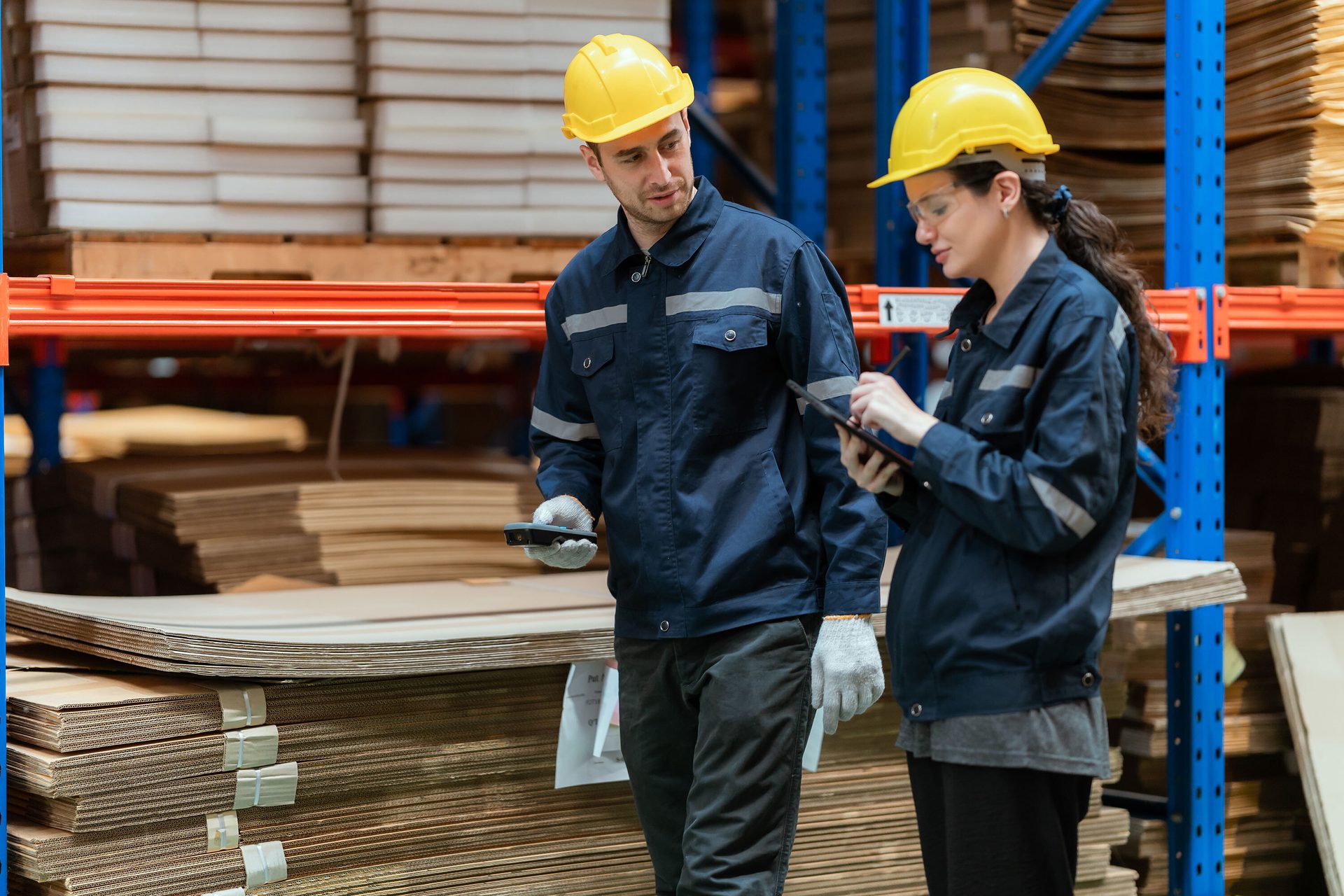 Two warehouse workers in safety gear, one with a tablet and one with a scanner, in a warehouse full of cardboard.