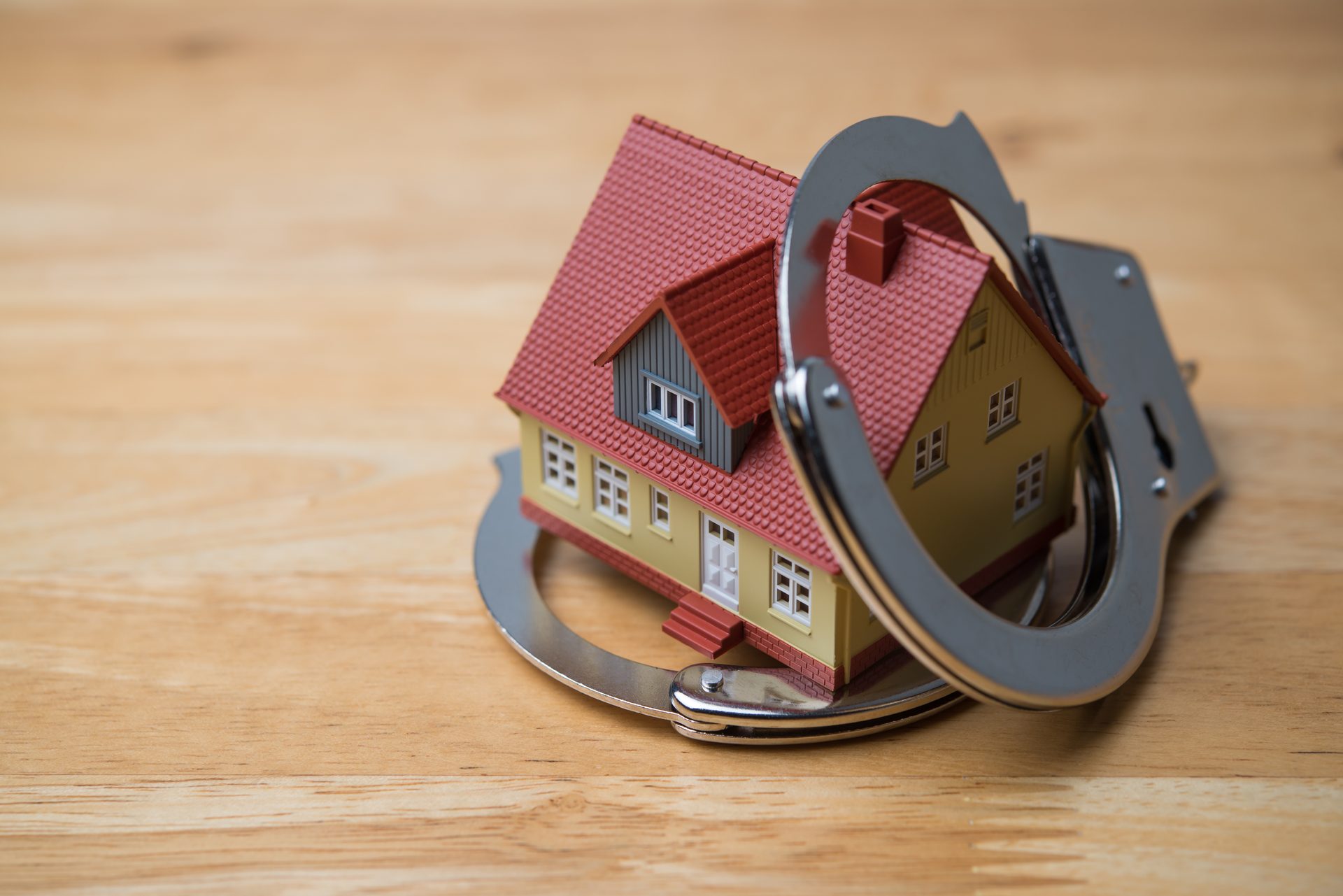 A miniature house model is shackled by metal handcuffs on a light wooden table, symbolizing property legal issues or debt.