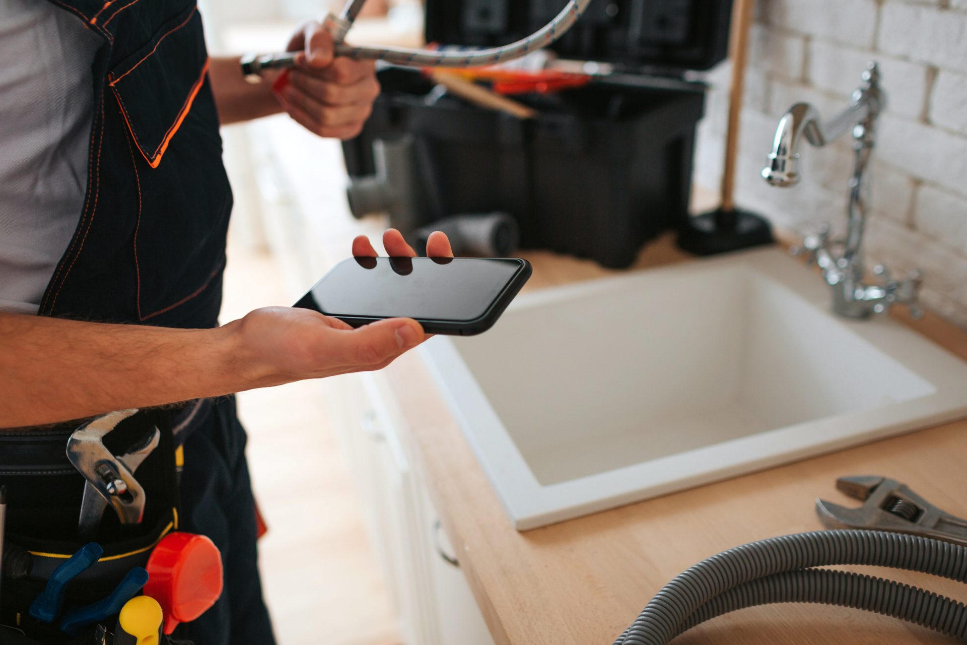 Plumber with smartphone and tools next to a kitchen sink, holding a hose, ready for repair work.