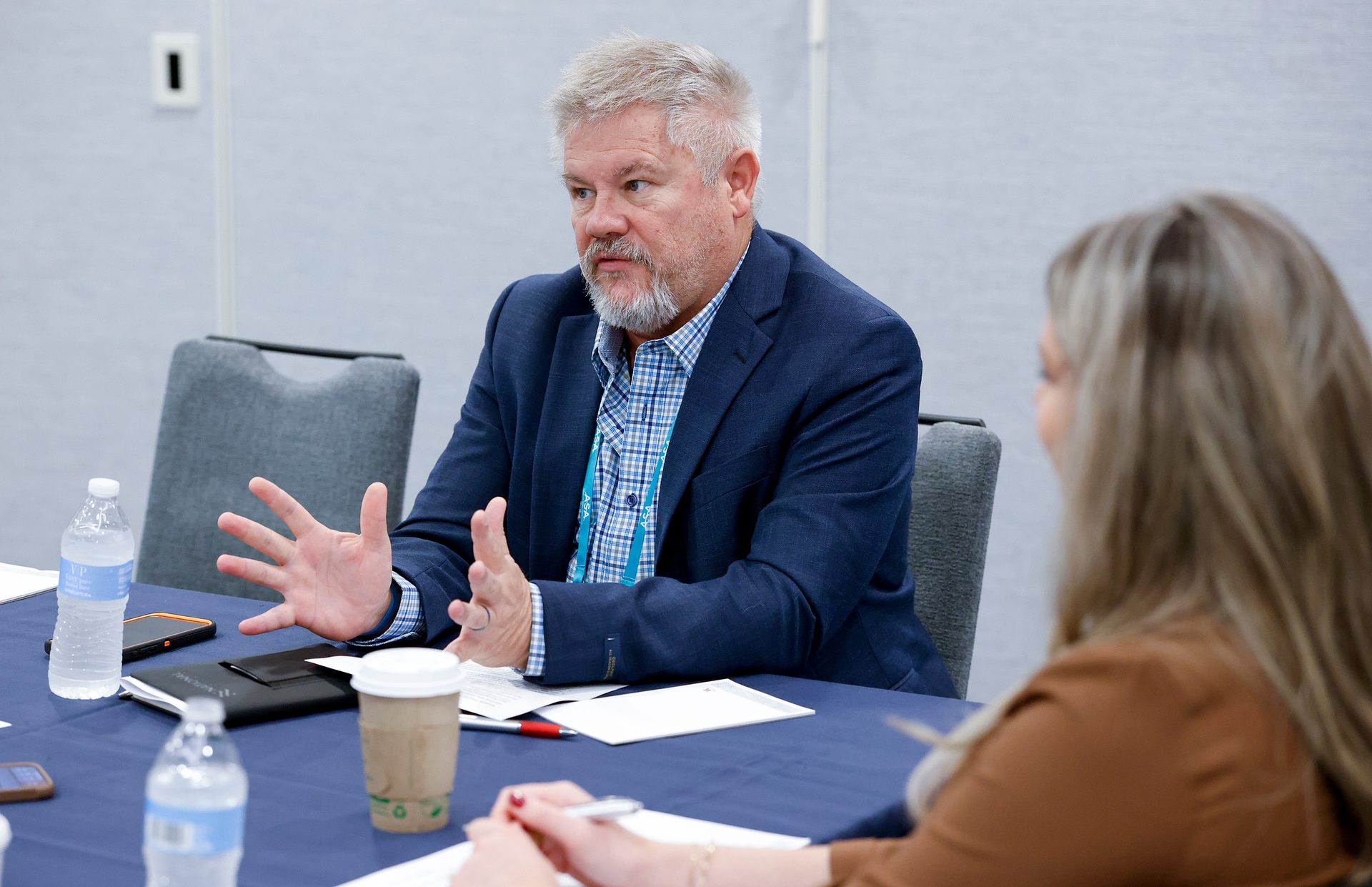 A bearded man in a blue suit jacket actively speaks and gestures at a table, facing a listener.