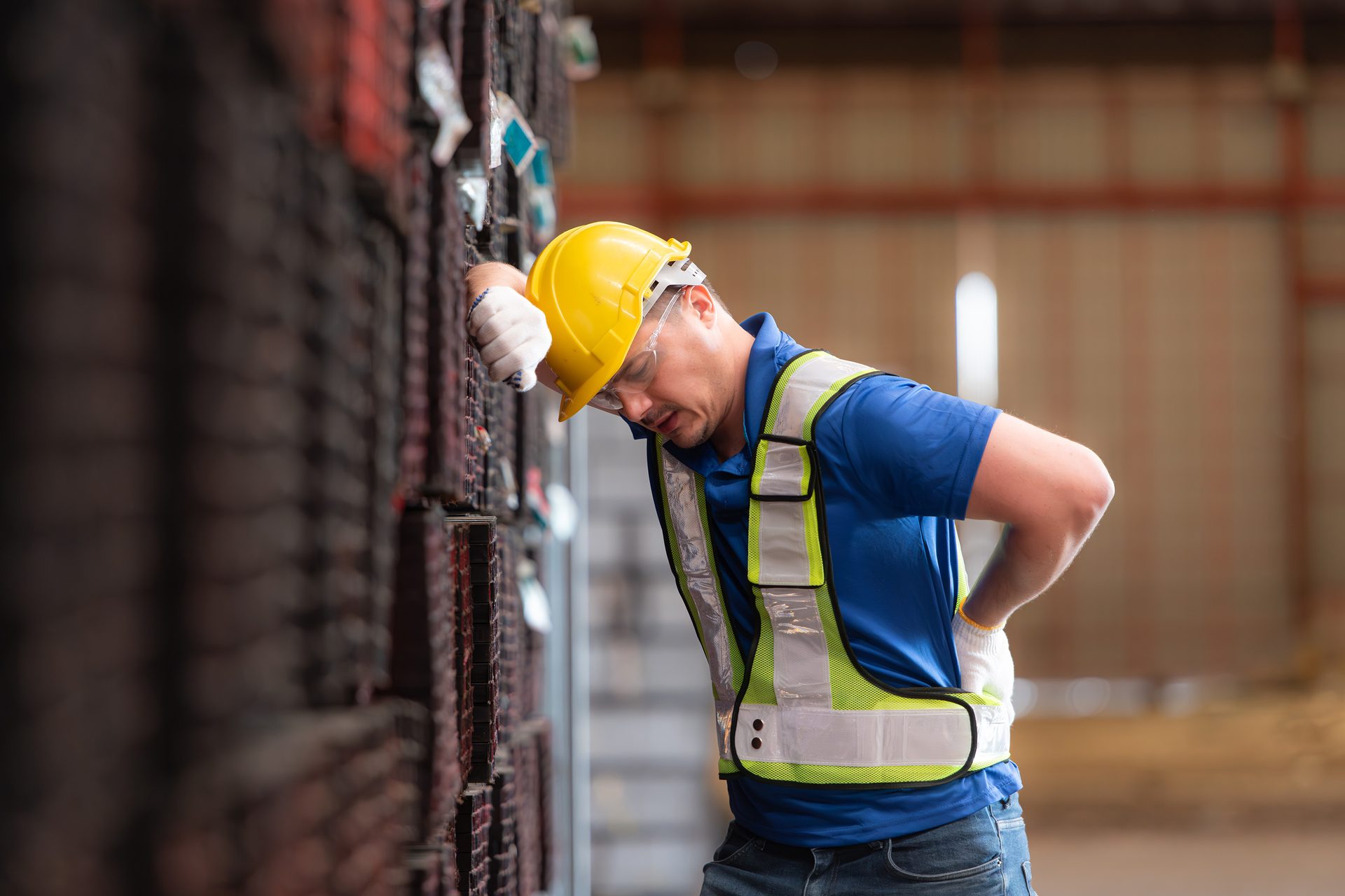 Factory worker in safety gear leans on shelves, clutching painful lower back.