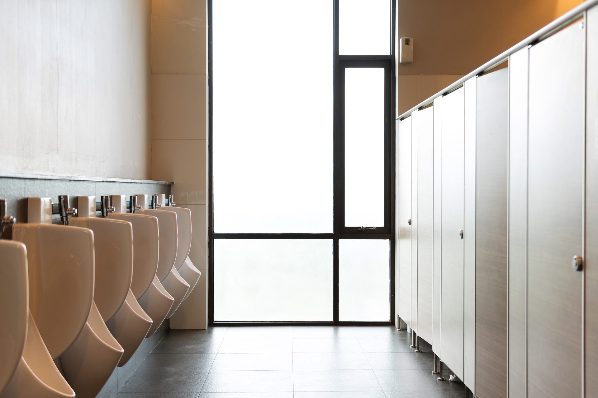 Clean men's restroom interior with a row of white urinals, large frosted window, and light-colored toilet stalls.
