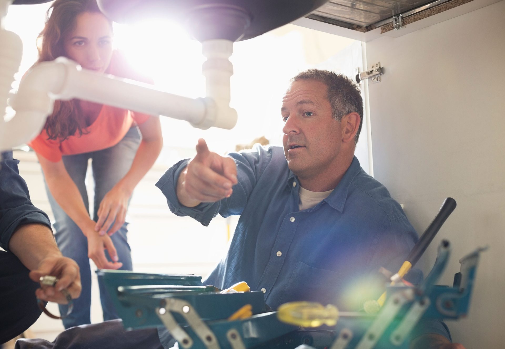 Man fixes plumbing under a sink, pointing, while a woman and another person watch.