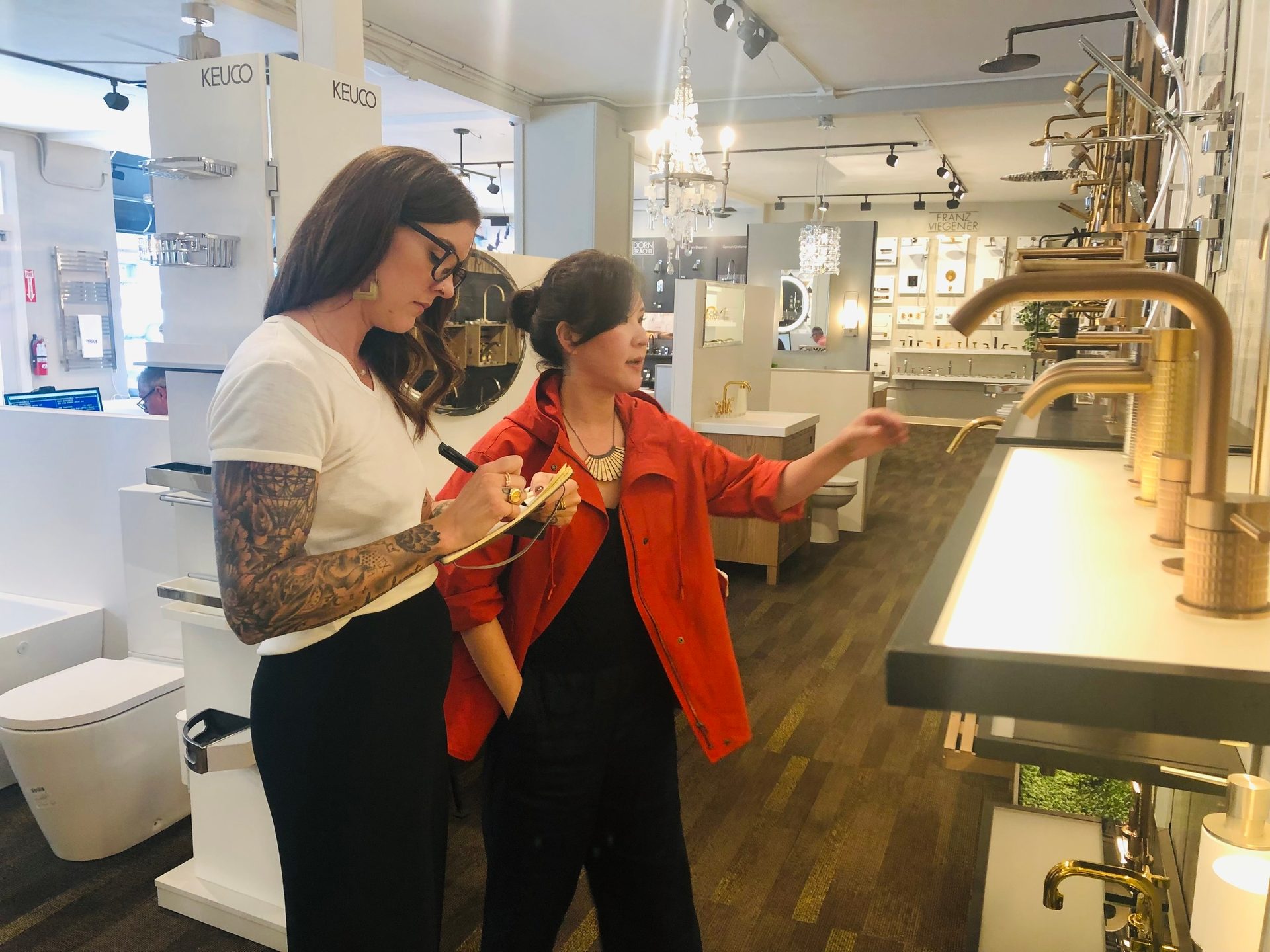 Two women consult at a plumbing showroom, one taking notes and the other pointing at brass faucets.