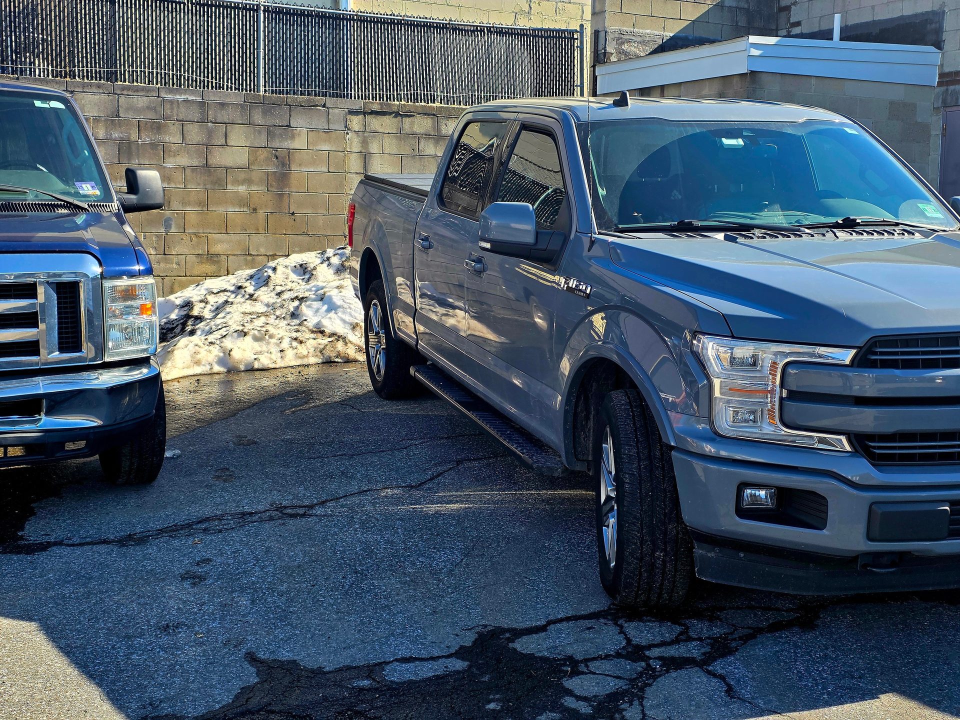 Two trucks parked on asphalt with a melting snow pile next to a brick wall and fence.
