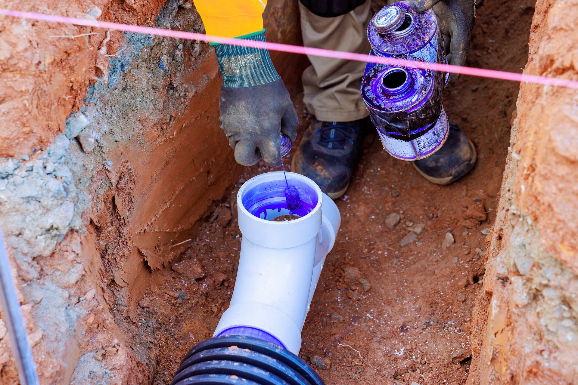 Person applies purple primer to PVC pipe in a trench.
