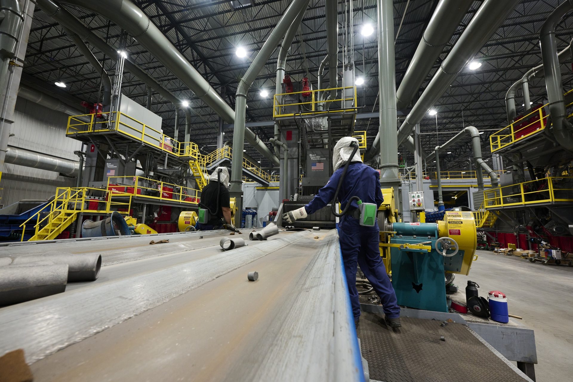 Workers in protective gear inspect pipe segments on a conveyor belt within a vast industrial facility.