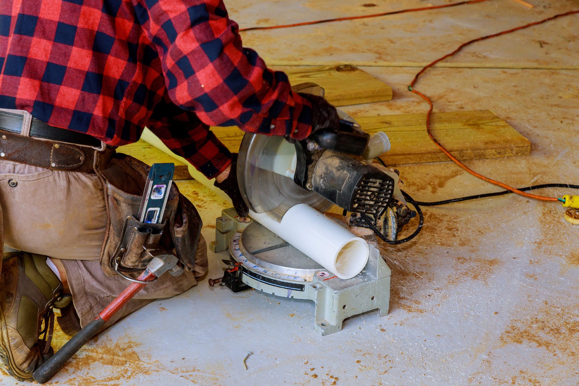 Worker in a plaid shirt cutting a white pipe with a miter saw, surrounded by sawdust.