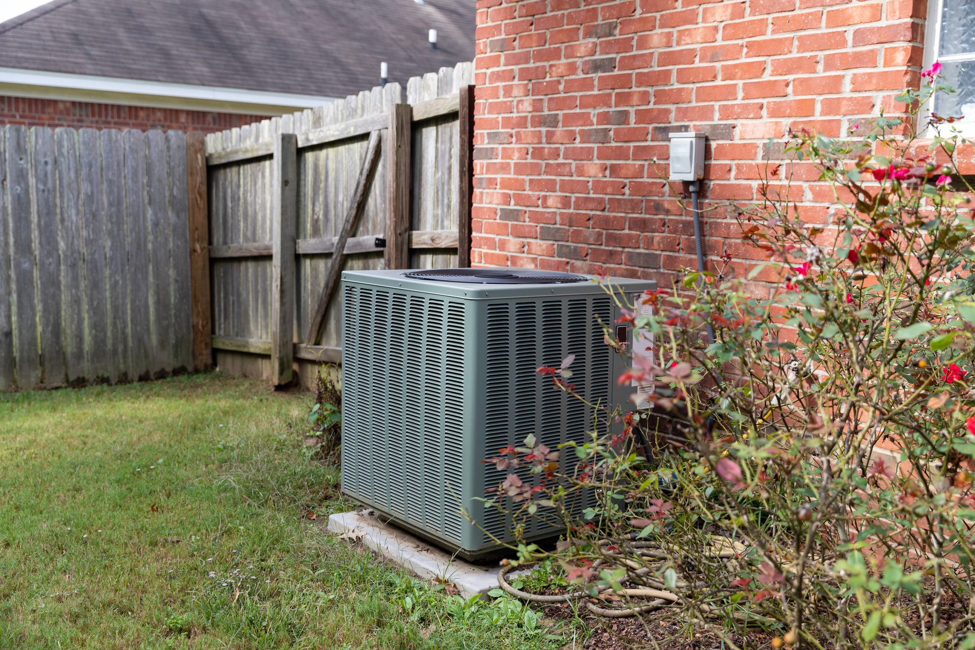 Outdoor AC unit beside a brick house and wooden fence, with a flowering bush.